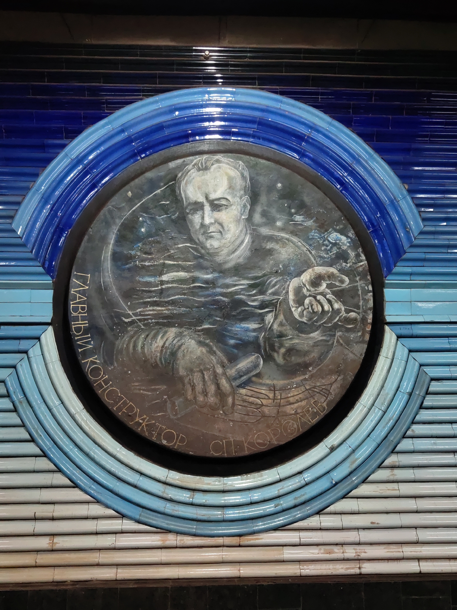 A circular mosaic depicts a man with short, dark hair and a serious expression, rendered in shades of gray and muted tones. He is wearing a dark shirt or jacket. His left hand is raised, holding a cylindrical object, while his right hand is extended, palm up, with fingers curled. The mosaic is set against a background of wavy, dark lines that suggest water or smoke, with faint specks of lighter color, possibly stars or debris. Around the circular mosaic, there is an arched frame made of layered, iridescent blue tiles. The mosaic is mounted on a wall clad in horizontal rows of light blue and white tiles, with a darker tiled border at the bottom. The overall impression is of a public artwork, likely a relief or mural. This artwork is located at the "Cosmonauts" metro station in Tashkent, Uzbekistan, honoring Sergei Korolev, a prominent rocket engineer and spacecraft designer. The inscription on the mosaic reads "GLADVY CONSTRUCTOR S.P. KOROLEV" in Cyrillic, meaning "Chief Designer S.P. Korolev."