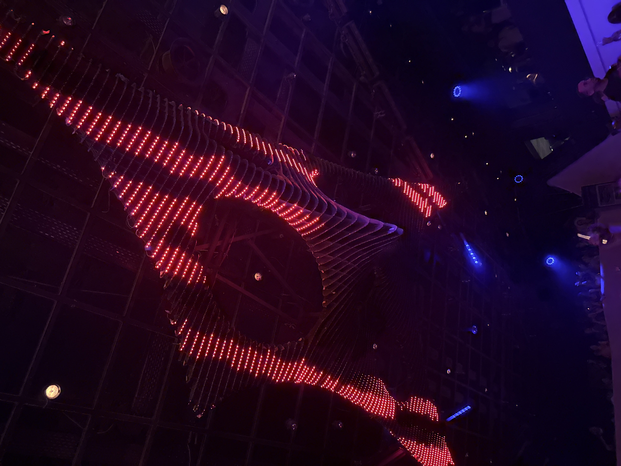 The image shows the interior of a dimly lit venue, possibly a nightclub or concert hall, with a focus on the ceiling and lighting. A large, undulating structure made of red LED lights dominates the upper portion of the frame, creating a vibrant, almost wave-like pattern. The ceiling itself appears to be a dark, possibly metallic, grid structure with some visible ventilation or structural elements.
Below the red LED installation, the space opens up into a darker area where blue lights are visible. These appear to be spotlights or accent lighting, casting beams into the darkness and illuminating parts of the crowd below. Silhouettes and indistinct figures of people are visible in the lower half of the image, suggesting an audience or patrons gathered in the space.
The overall atmosphere is one of a lively event, characterized by dynamic lighting and a sense of energetic gathering. The exact location in Mykonos, Greece, cannot be determined from the visual information alone, as the image focuses on the internal structure and lighting of the venue rather than any external or identifiable landmarks.