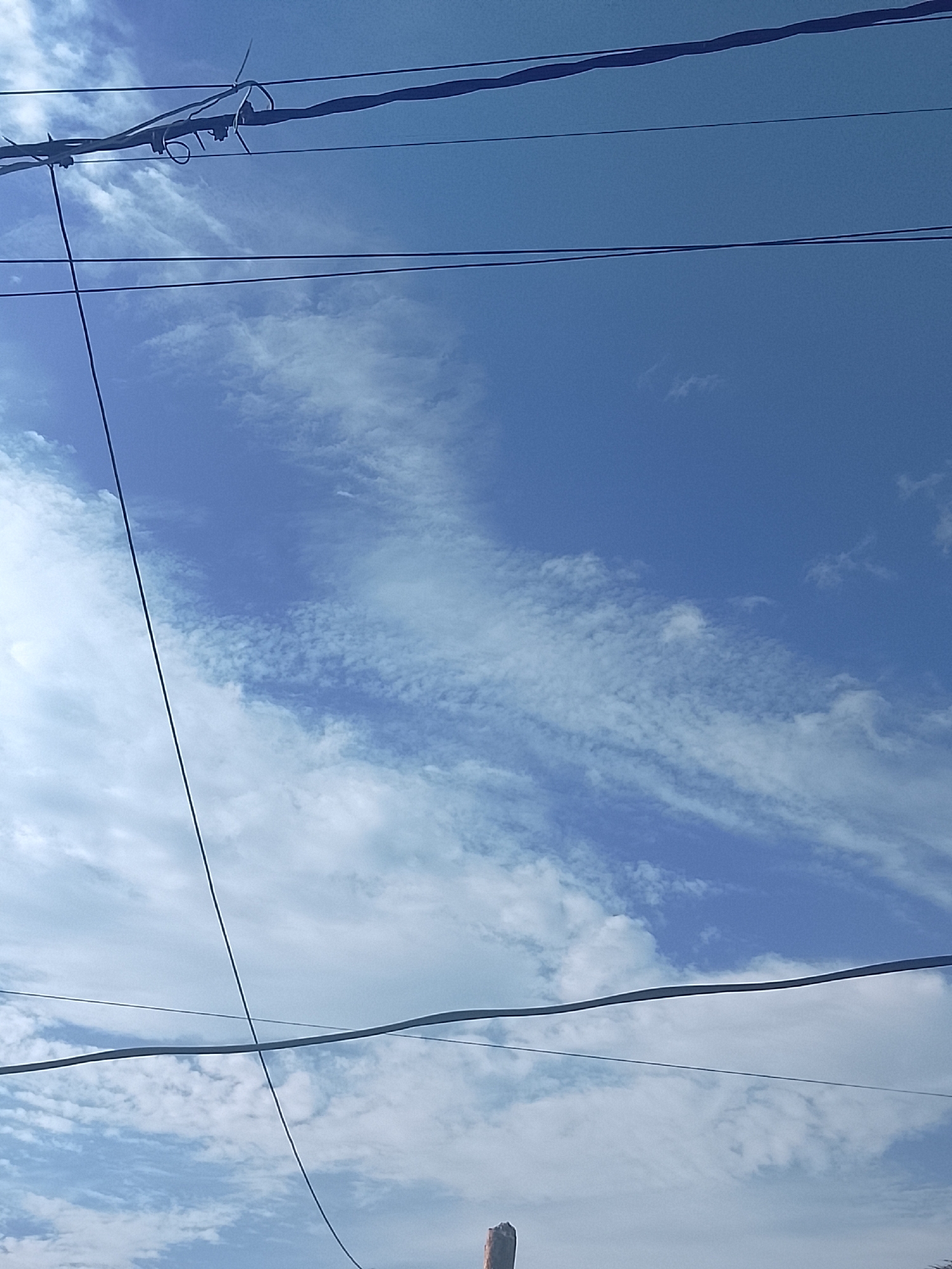 A low-angle shot taken in Altagracia, Venezuela shows a blue sky with scattered clouds. Several power lines cut across the frame. The top of a wooden pole is visible near the bottom center of the image.