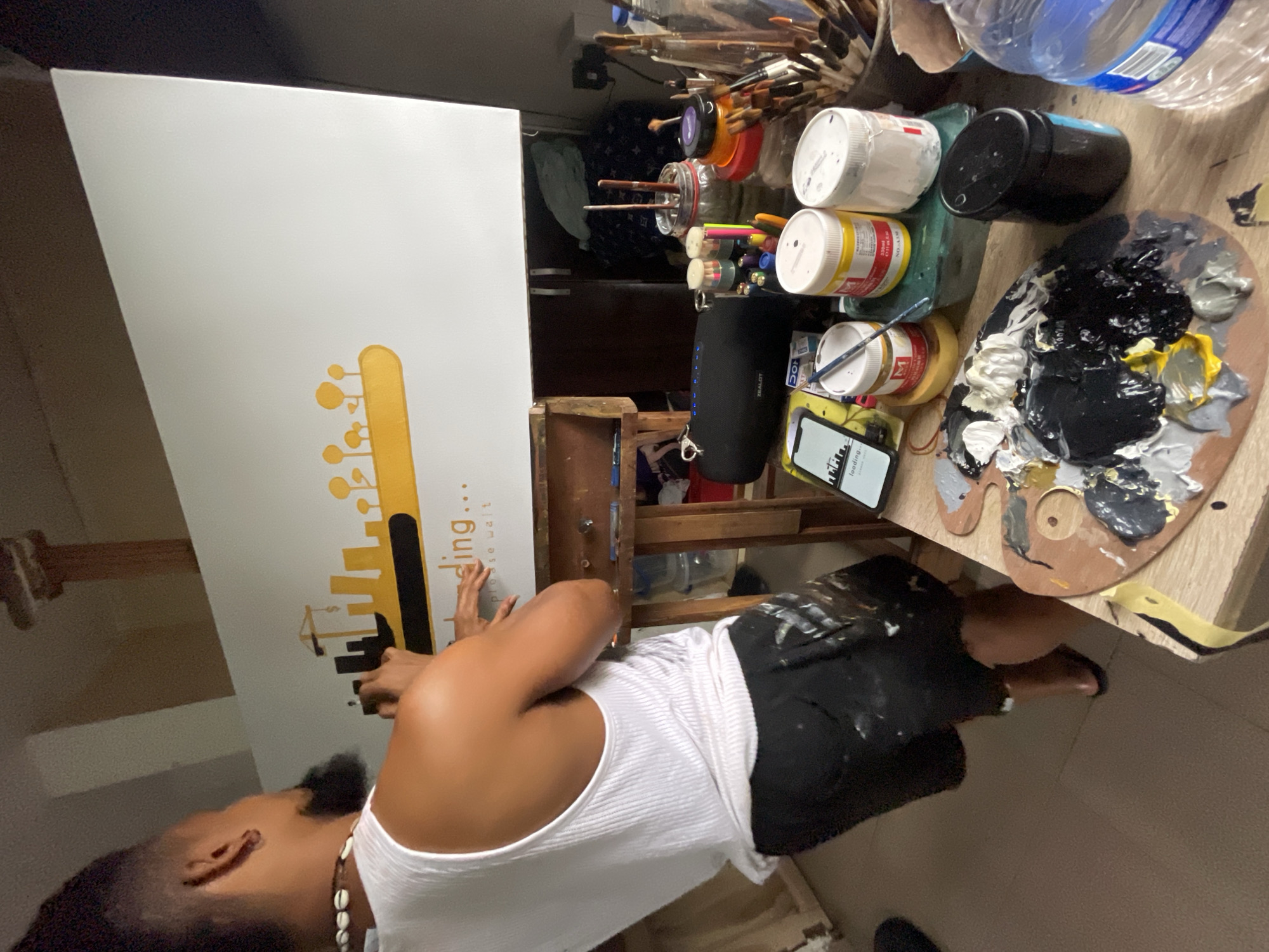 A man is painting in a room. The room appears to be in Ado, Nigeria. The man is wearing a white tank top, black shorts with paint stains, and a necklace of seashells. He stands facing a canvas on an easel. The canvas features a yellow silhouette of a city skyline with a stylized guitar neck above it. Some text in lowercase letters appears beside the illustration.
To the right of the easel, there is a table cluttered with paintbrushes, jars of paint, and a wooden artist's palette covered in dabs of black, white, gray, and yellow paint. There's also a black cylinder-shaped speaker and a smartphone. A bottle of water is among the items on the table. In the background, a dark cabinet is visible.