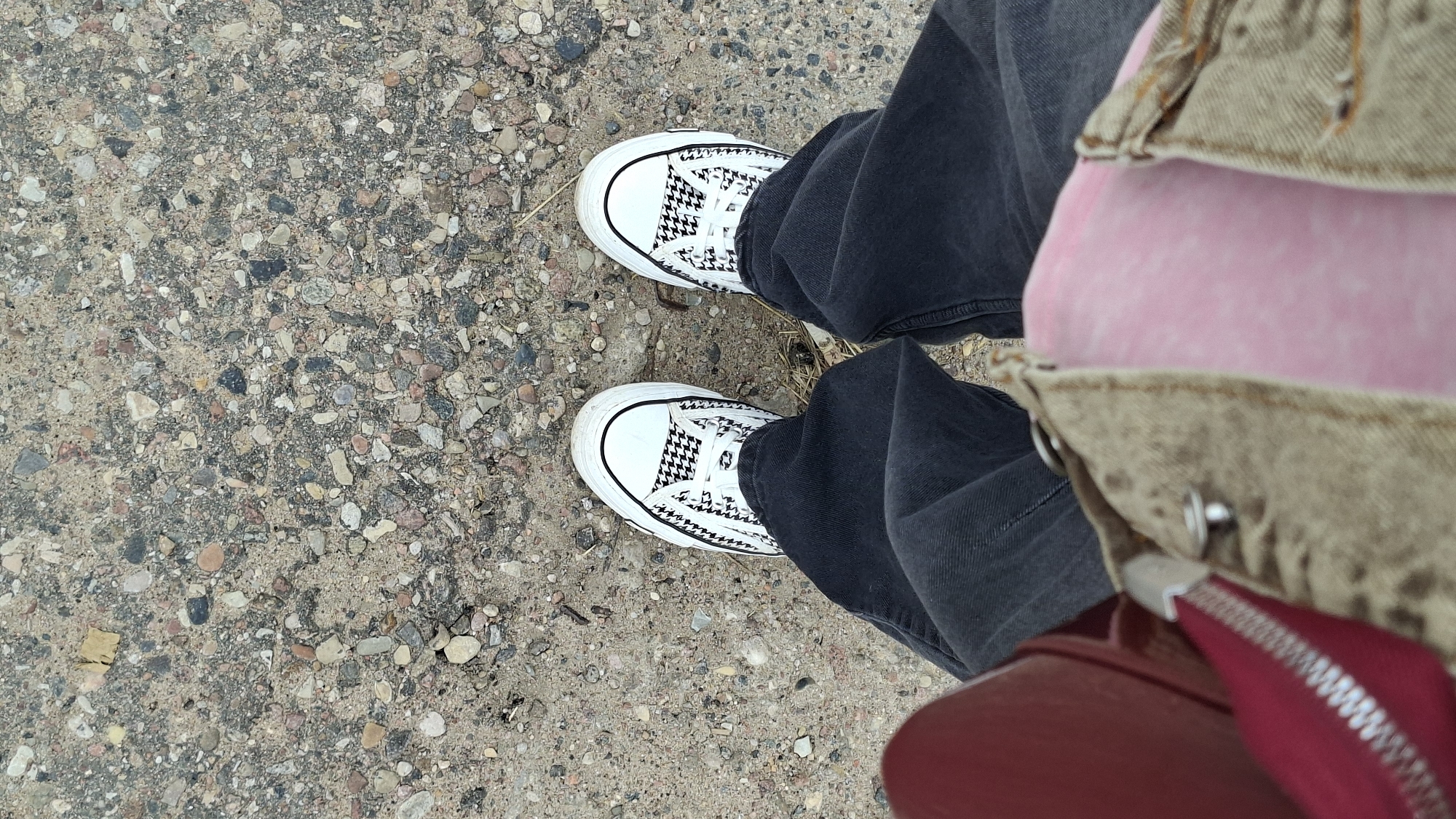 The image shows a pair of white canvas sneakers with a black houndstooth pattern on a dirt and gravel ground. The sneakers are laced with white laces. The person wearing the shoes has on dark gray jeans. Part of a tan denim jacket or vest is visible, as is a pink object and a red bag with a silver zipper. The scene appears to be outdoors, possibly in a rural area given the ground cover. The lighting suggests it is daytime. There is no visible text in the image. The setting is identified as Забалоцкі сельскі Савет, Belarus.