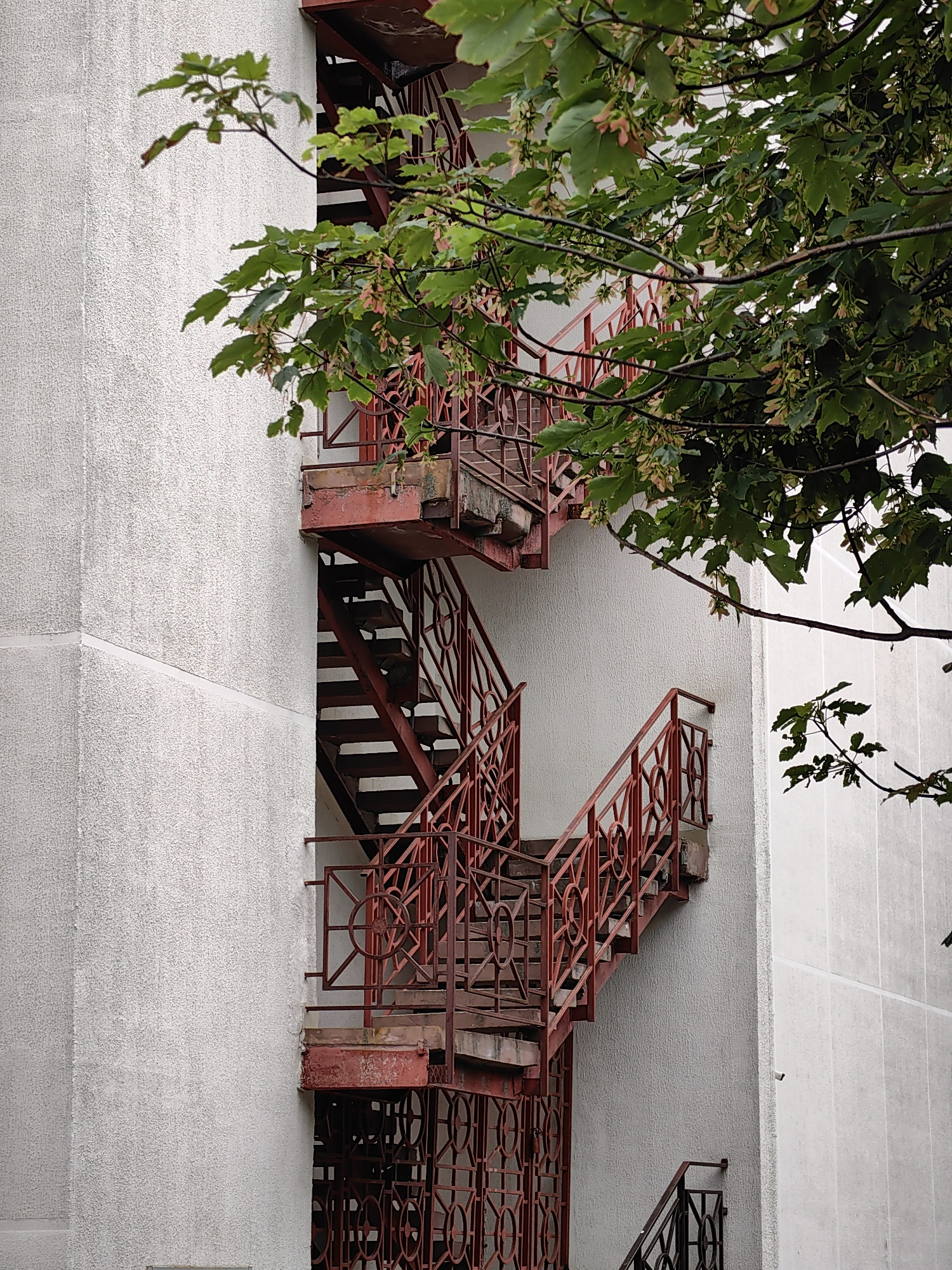 A red metal fire escape is visible on the side of a white, textured building in Minsk, Belarus. The fire escape features multiple flights of stairs with decorative metal railings. The stairs are made of metal grates. Overhanging the fire escape and the building is a tree with green leaves and some seed pods. The sky appears to be overcast, contributing to the muted lighting of the scene. There are no people visible in the image.
