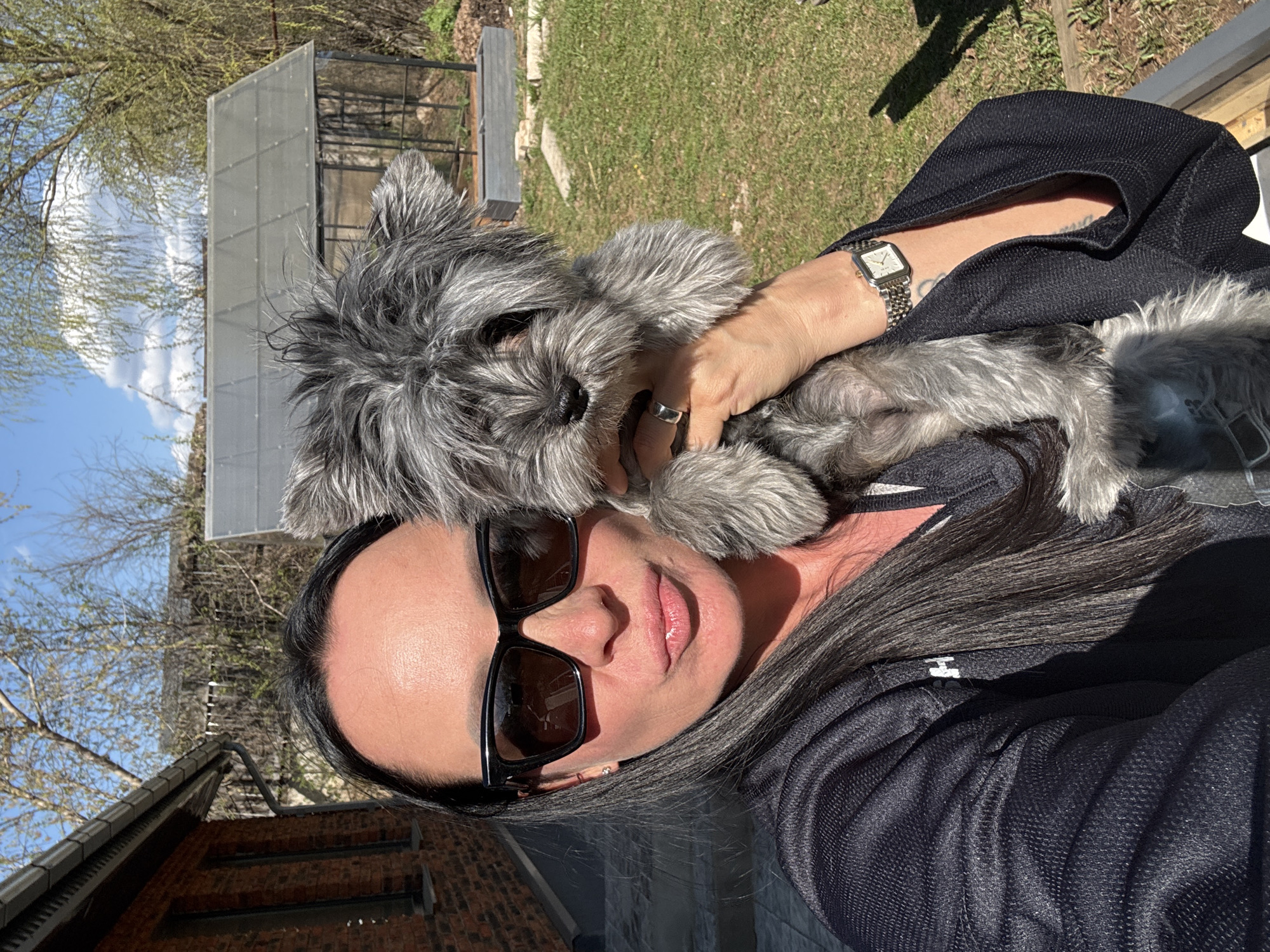 A middle-aged woman with long, gray hair, wearing sunglasses and a dark jacket, holds a gray, wire-haired dog in her arms. She is wearing a watch and a ring. They are outdoors; the background includes a brick building, a greenhouse, trees, and grass. The location is likely in Chaikovsky, Russia.