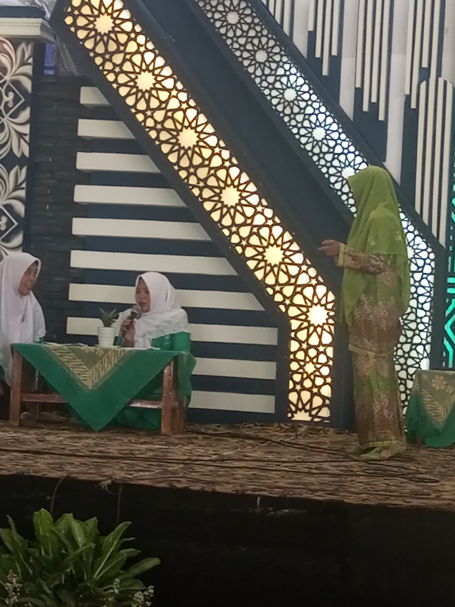 Three individuals are on a raised wooden platform in what appears to be an indoor venue in Klaseman, Indonesia. Two women are seated at a table covered with a green cloth, and a third woman stands to the right, facing them. The woman on the left wears a light-colored hijab and a matching outfit. The seated woman on the right wears a white hijab and a green outfit, holding a microphone and speaking. The standing woman to the right wears a light green hijab and a patterned green and brown outfit. Behind the individuals, a backdrop features decorative, illuminated geometric patterns and dark, striped panels. In the foreground, lush green foliage is visible.