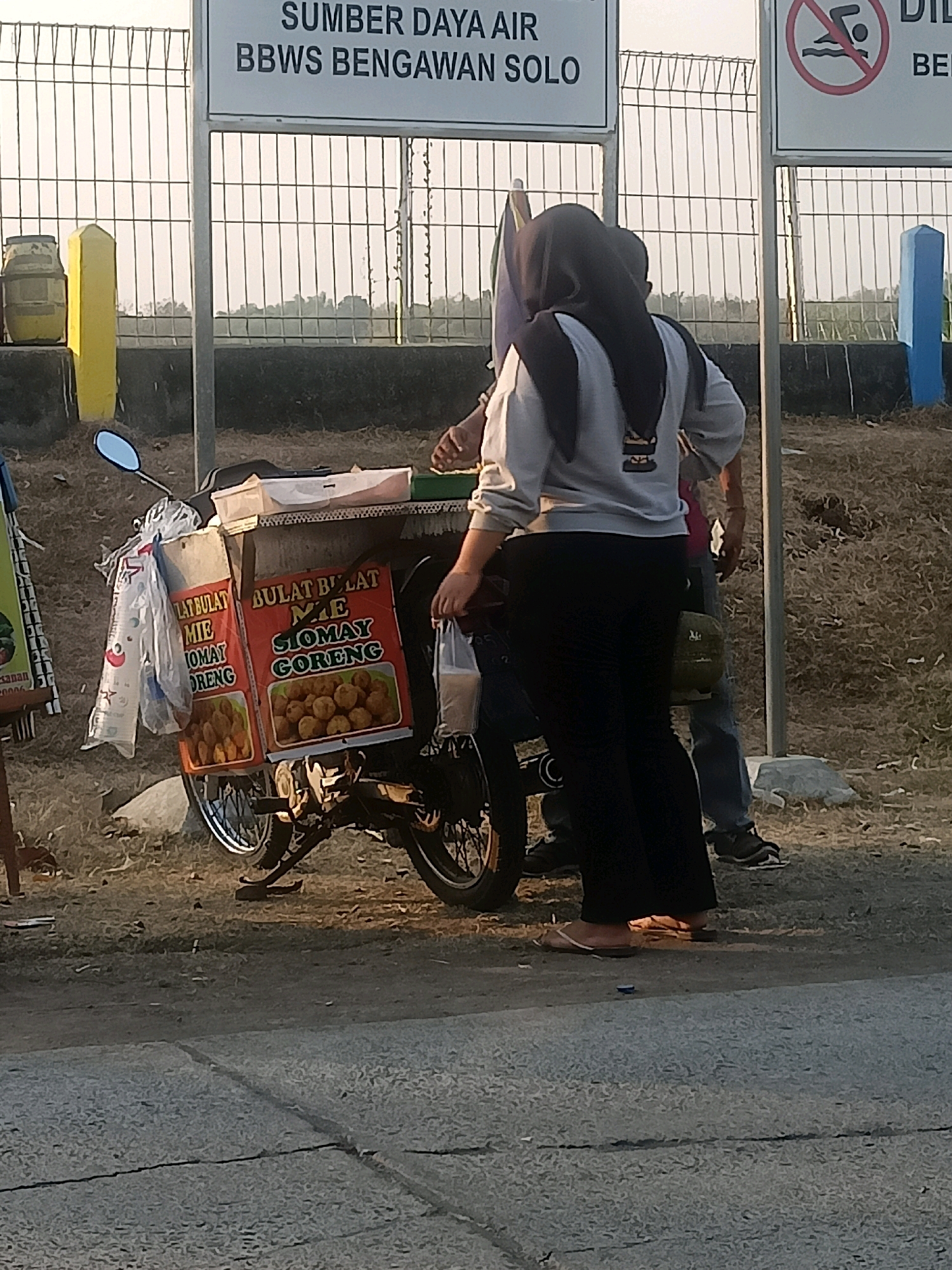 The image shows a street scene in Ketro, Indonesia, where a motorcycle is equipped with a food cart. The cart displays signage for "Siomay Goreng" (fried siomay), a popular Indonesian snack. Two people are standing near the cart. One person, wearing a hijab and black pants, is standing with their back to the camera, holding a plastic bag. Another person, wearing a blue shirt and jeans, is partially visible behind them. The background shows a metal fence and dry vegetation. A sign above the fence reads "SUMBER DAYA AIR BBWS BENGAWAN SOLO," indicating the presence of a water resource management agency. Another sign with a "no swimming" symbol is also visible. The overall scene suggests a commercial street activity in the vicinity of a water-related facility.