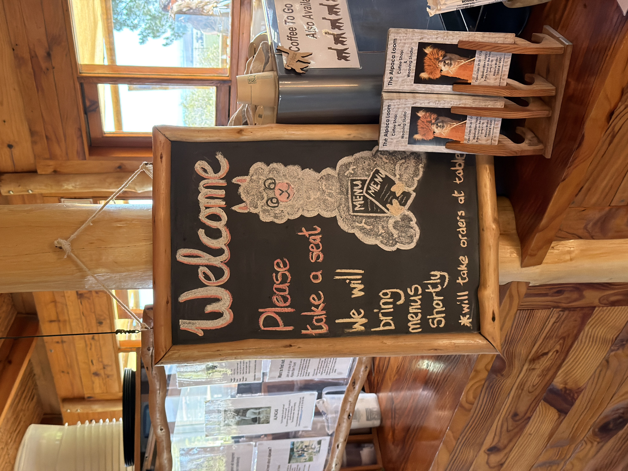 This media file captures the inviting interior of a rustic establishment, likely a coffee shop or weaving studio, characterized by extensive use of natural wood for walls, furniture, and window frames.
The central subject is a black chalkboard sign, edged with natural wood, prominently displaying a "Welcome" message in red and white chalk. Below, the sign instructs: "Please take a seat We will bring menus Shortly *will take orders at table." A charming, fluffy alpaca character, drawn in chalk, holds a "MENU" sign, reinforcing the business's theme.
To the right of the chalkboard, a wooden display stand holds several brochures titled "The Alpaca Loom Coffee Shop & Weaving Studio." These brochures advertise "Open Daily 09:30 - 17:30," "Alpaca Barn Tours Every hour on the hour," and "Alpaca Walks: 10:00 & 16:00." Further right and slightly behind, another sign indicates "Coffee To Go Also Available" and features silhouettes of alpacas.
In the background, large wooden-framed windows reveal bright daylight and glimpses of green trees, suggesting a clear, sunny day. The overall setting is warm and natural, implying a tranquil, welcoming atmosphere. The visible text clearly indicates that the establishment is "The Alpaca Loom," offering coffee, a weaving studio, and alpaca-related experiences. This fits the location context of Drakenstein Nu, South Africa, suggesting a farm or nature-based visitor attraction.