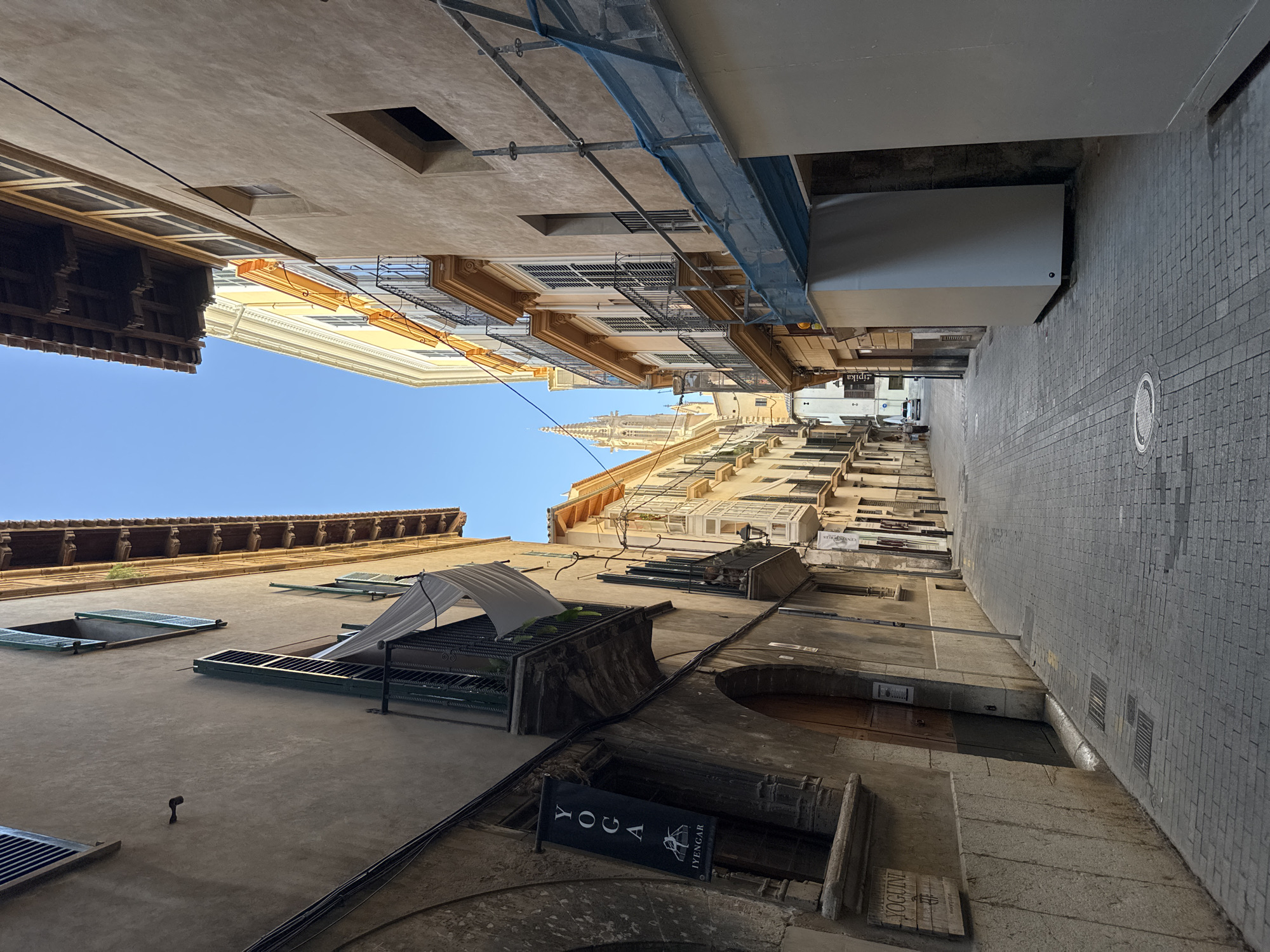 The image shows a narrow, paved street in Palma de Mallorca, Spain, lined with tall, old buildings. The street slopes gently upwards towards a distant church spire visible between the buildings. The buildings on either side are made of stone and plaster, with traditional wooden shutters on the windows and balconies. Some buildings are under construction or renovation, indicated by scaffolding and blue netting.
On the left side of the street, a dark banner hangs from a building, displaying "YOGA" in white lettering above "IYENGAR" and a silhouette of a yoga pose. Below this banner, a wooden sign reads "YOGUINI".
On the right side of the street, there are several white structures that appear to be temporary enclosures or building materials. The street is paved with cobblestones. The sky is clear and blue. There are no people prominently featured in the foreground, but two figures are visible in the distance walking down the street. The overall impression is of a historic European city street in sunlight.