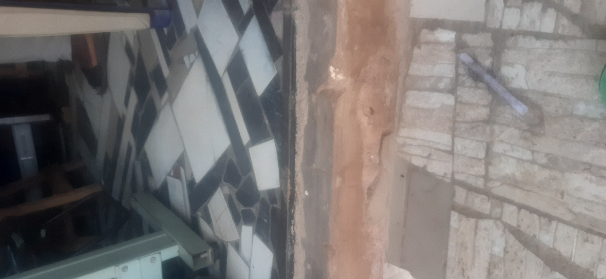 The image shows a close-up, downward-angled view of a tiled floor in Jos, Nigeria. The floor is covered with broken, irregularly shaped tiles, some black and some white, arranged in a chaotic pattern. A section of the floor shows a different type of paving, consisting of rough, rectangular stones laid close together. In the upper portion of the image, part of a white metal structure, possibly furniture or construction material, is visible. The scene appears to be outdoors or in an open, unfinished area, given the rough texture of the stone paving and the presence of what looks like disturbed earth or debris. There are no people clearly visible in the frame, although a sliver of a person's leg wearing dark footwear is present in the upper right quadrant. A single, thin, light-colored object, possibly a piece of paper or plastic, lies on the stone paving in the lower right quadrant.
