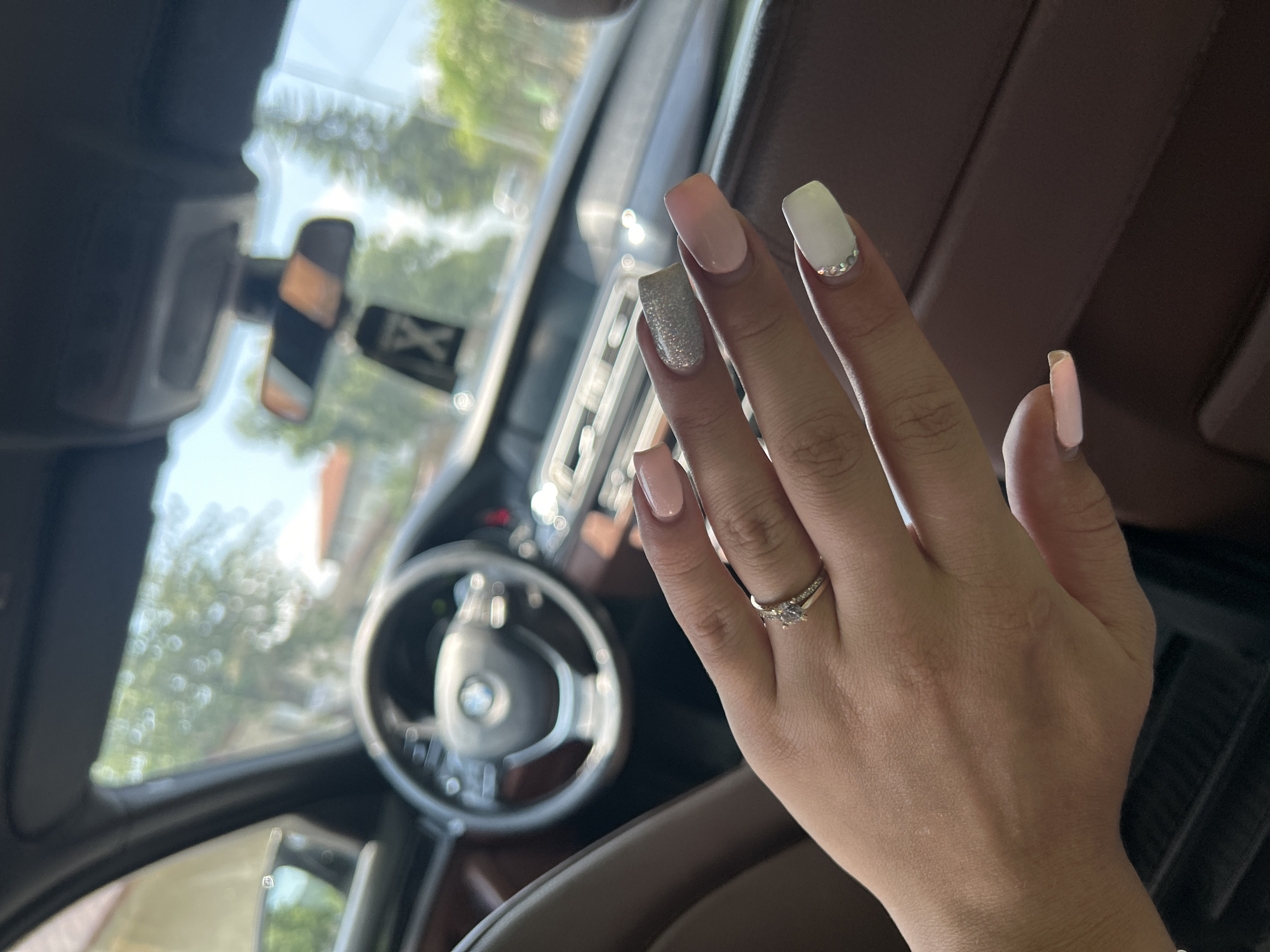 The image displays a person's right hand, adorned with a manicure and rings, positioned inside a car with a bright outdoor background.
**Subjects:** The primary subject is a human right hand. Elements of a car interior and blurred exterior scenery are secondary subjects.
**Objects:** The hand features square-shaped nails painted in a varied style: pale pink on the pinky and middle fingers, sparkling silver glitter on the ring finger, and off-white with a line of small rhinestones near the cuticle on the index finger. On the ring finger, a prominent ring with a central stone flanked by smaller stones is worn, accompanied by a thinner band underneath it. The car's steering wheel, clearly displaying the BMW logo, is in the blurred mid-ground. Parts of the dashboard, a rear-view mirror, and a side mirror are also visible.
**Scene/Setting:** The scene is set inside a modern car, identified as a BMW. The car appears to be stationary or moving slowly, allowing for a clear focus on the hand. Through the car windows, a bright, sunny outdoor environment with blurred green trees and foliage is visible. The provided location context is Madara, Bulgaria.
**Activity or event taking place:** The image captures the casual display of a recent manicure and a ring, likely a personal snapshot taken while traveling or waiting in the car.
**Notable details:** The photo is taken in bright daylight, indicating a sunny day. The manicure exhibits a mix of subtle and glittery nail polish, with decorative rhinestones. The focus is sharp on the hand, while the background is intentionally blurred to highlight the subject.
**Visible text:** The "BMW" logo is clearly identifiable on the steering wheel. A faint, blurry white "X" on a dark background is discernible in the reflection within the rear-view mirror.