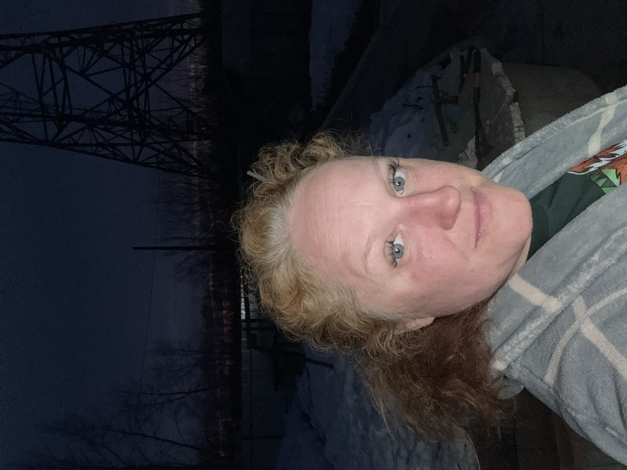 A selfie captured in Chaikovsky, Russia, at dusk or night, shows a middle-aged woman with fair skin, blue eyes, and curly reddish-blonde hair, wearing a patterned grey robe over a dark green shirt. The background includes a tall power line structure and a partially snow-covered area with a concrete structure visible. The darkness obscures details of the surrounding environment.