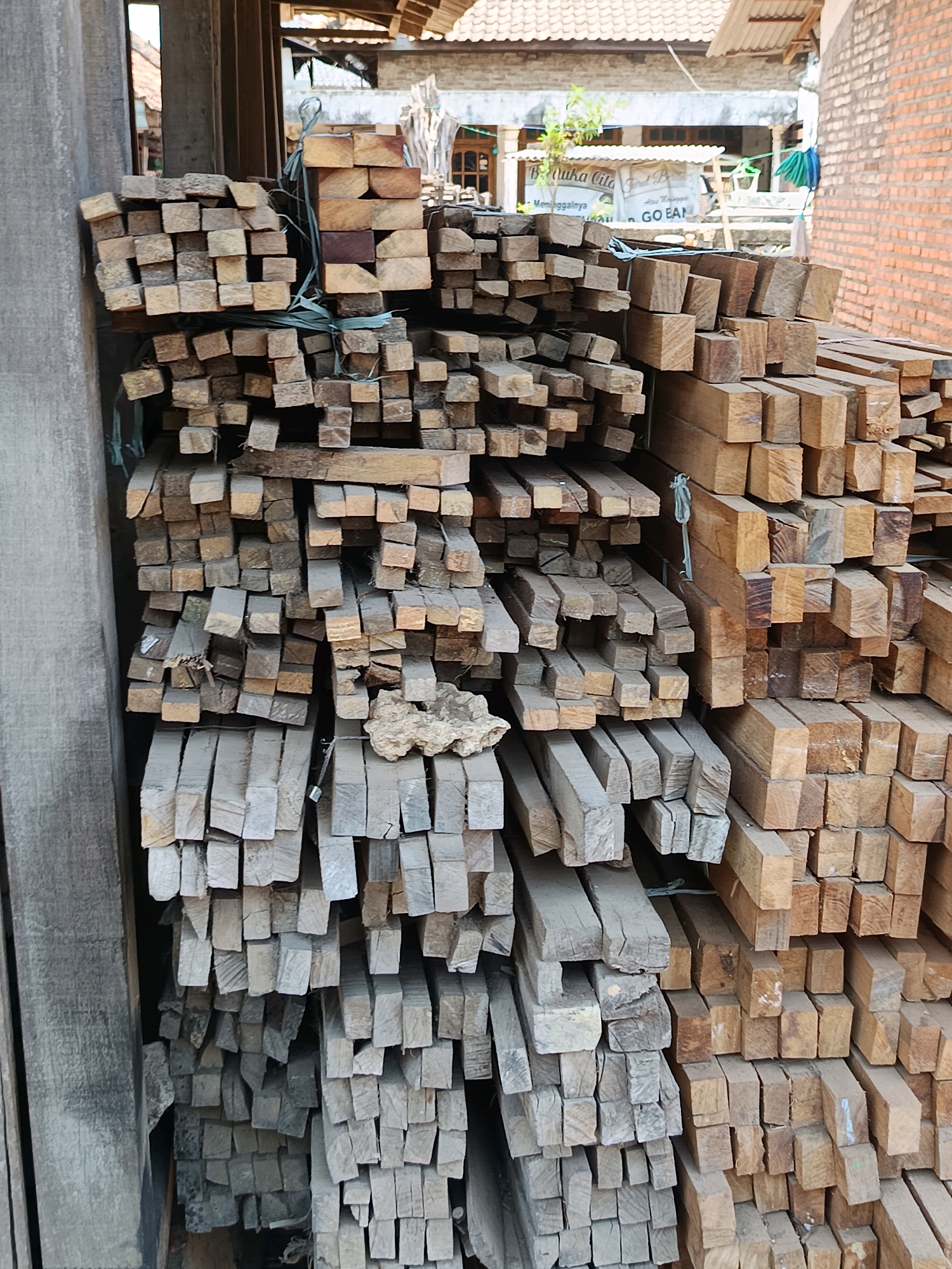 The image shows a large stack of cut lumber, predominantly square in cross-section, with some rectangular pieces visible. The wood appears to be unfinished and is stacked in an outdoor setting. In the background, there is a brick wall on the right and a building with a tiled roof and what looks like a banner or sign on the left. The overall impression is that of a lumber yard or a construction site. The location is identified as Blega, Indonesia.