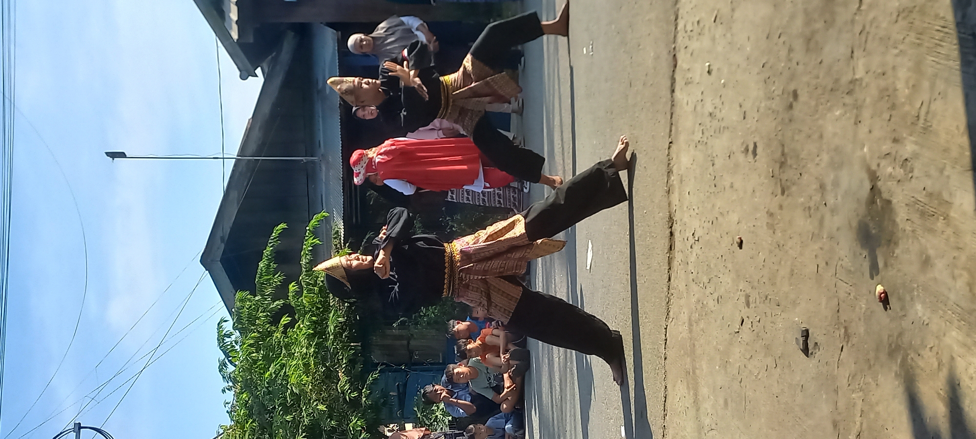 The image depicts an outdoor cultural performance in Samarinda, Indonesia. Two individuals, likely performers, are engaged in a martial art or dance, dressed in traditional attire consisting of black tops, head coverings, and ornate sashes or wraps around their waists and hips, paired with dark, loose-fitting pants. They are positioned in a dynamic stance, with their arms raised and bodies bent.
Behind the performers, a crowd of spectators, including children and adults, are seated or standing, observing the event. Among the spectators, a person wearing a red cloak and hat stands out. The background features residential buildings with dark, sloped roofs and trees. The ground appears to be a paved or dirt road with some debris scattered on it. Overhead, utility wires stretch across the sky, indicating an urban or semi-urban setting. The lighting suggests it is daytime, with a clear sky.