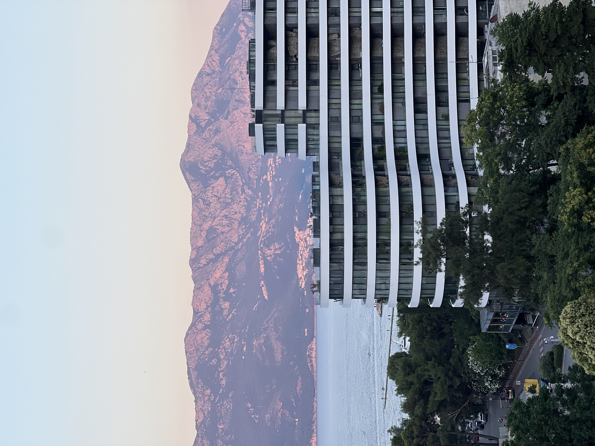 The image depicts a scenic view of Budva, Montenegro, at sunset. In the foreground, a modern, curved white building with multiple balconies and large windows dominates the right side of the frame. Below this building, lush green trees and vegetation line a street with parked cars and a few pedestrians. The street appears to be a city road with traffic lights.
The middle ground features a serene body of water, likely the Adriatic Sea, with a pier extending into it. Several small boats are visible on the water, and a few individuals are seen on the pier and near the water's edge.
In the background, a majestic mountain range is illuminated by the warm hues of the setting sun, casting a soft pink and orange glow on its slopes. The mountains are dotted with small clusters of buildings, suggesting the presence of villages or settlements along the coastline. The sky above transitions from a pale blue at the top to a soft pinkish-orange near the horizon. The overall atmosphere is tranquil and picturesque, showcasing the natural beauty of Budva and its surrounding landscape.