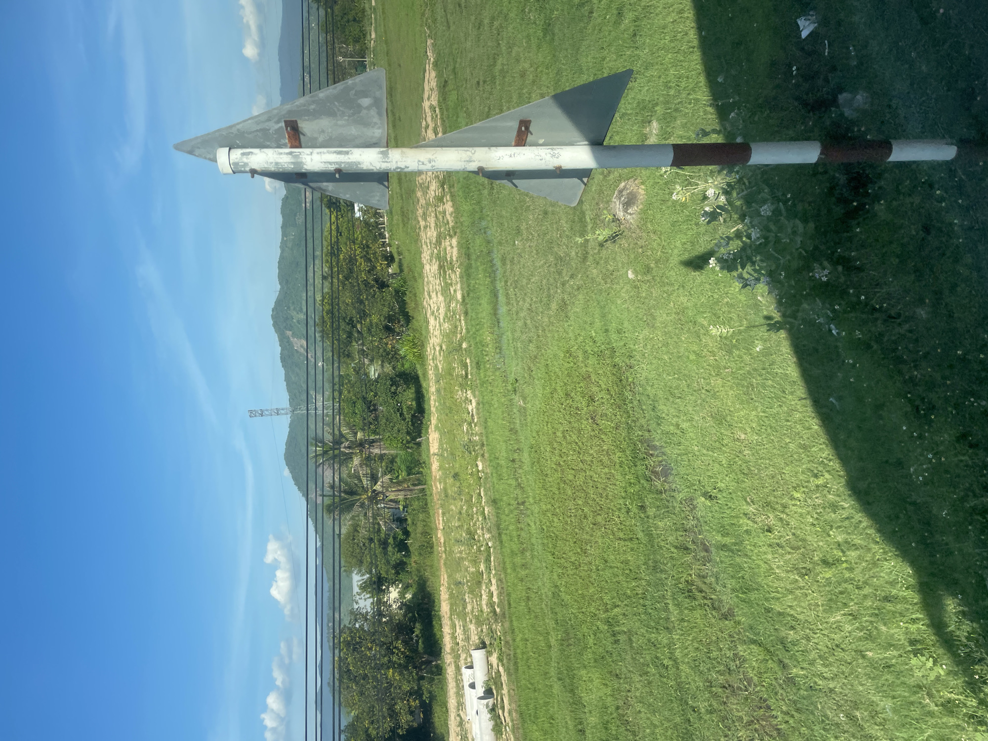 A roadside view in Tay Nha Trang Ward, Vietnam, shows a tall pole with two triangular gray signs, one above the other. The pole is painted with alternating red and white bands. In the foreground, a grassy field is visible, with some patches of wet ground and small white flowers. In the background, there are trees, a distant mountain range under a clear blue sky with scattered clouds, and overhead power lines with a transmission tower. There are also some concrete pipes lying on the ground to the left of the frame. No people are present.