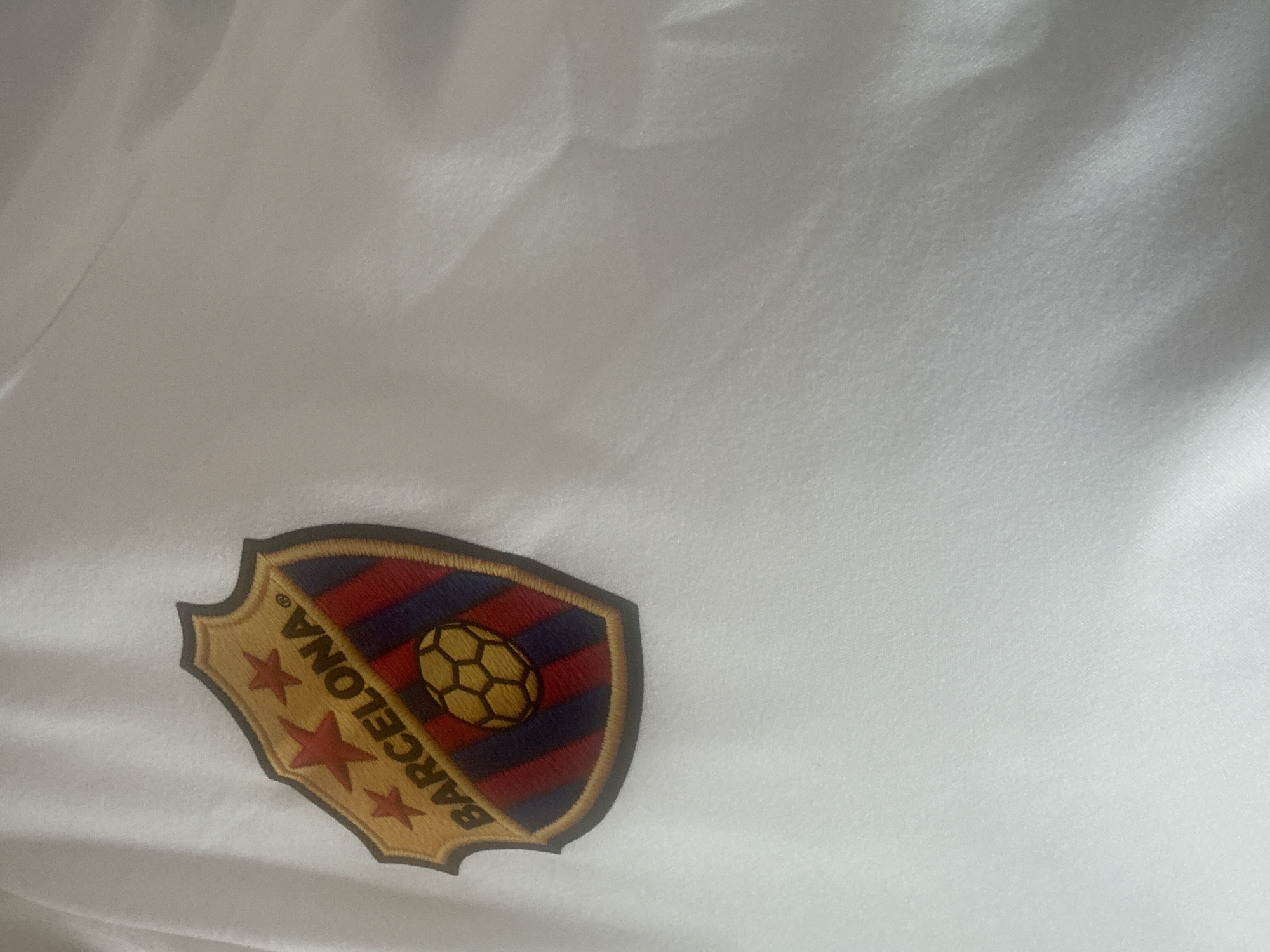 An embroidered shield-shaped patch is affixed to a white textile surface in the city of Unknown, Unknown. The patch features a gold-toned border. Its upper section displays a gold background with three prominent red five-pointed stars. Below this, the word "BARCELONA" is embroidered in dark capital letters, slightly arched, on a gold background, followed by a registered trademark symbol (®). The lower portion of the shield contains alternating vertical stripes of red and dark blue/purple, with a yellow and black outlined soccer ball centrally placed within this striped area. The white textile background exhibits subtle folds and shadows, indicating a soft, draped fabric, possibly a garment. The lighting appears uniform across the scene.