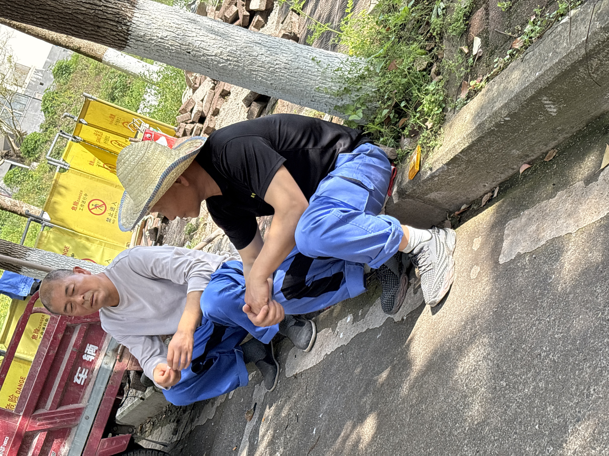 Two men are sitting close together on a concrete surface adjacent to a curb, with a red truck visible to the left. The man on the left is wearing a light gray long-sleeved shirt and blue pants, while the man on the right wears a black t-shirt, blue pants, and a straw hat. They appear to be interacting, possibly holding hands. Yellow signs with safety warnings are visible in the background. A tree trunk lies horizontally near the top of the frame, with grass and brick rubble behind the curb. The location is identified as Jiangjin, China.