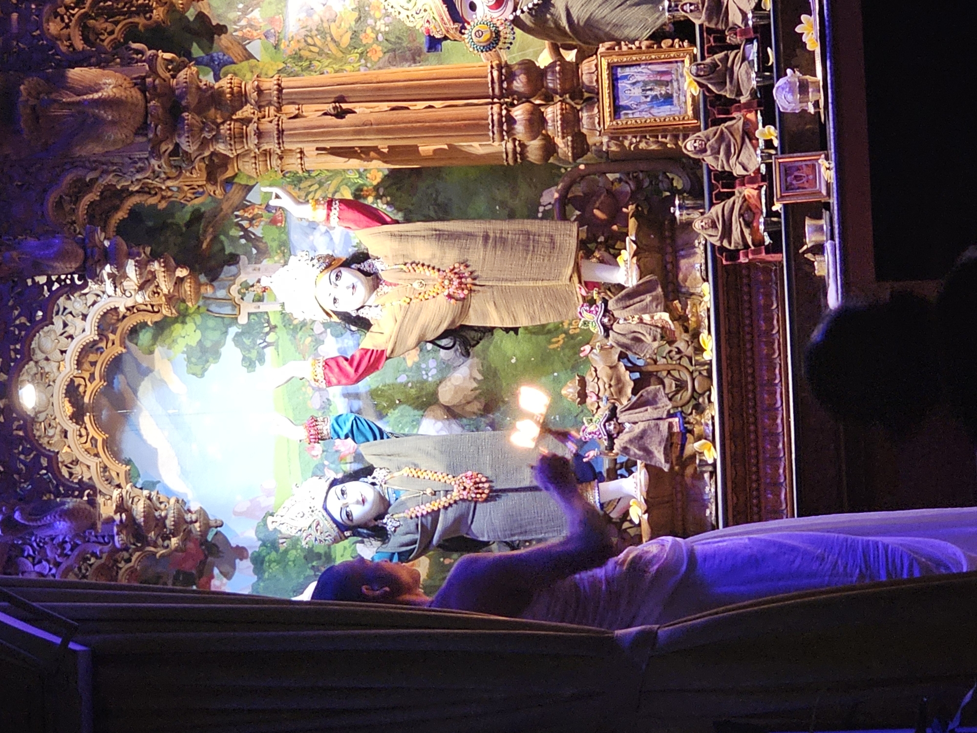 The image captures a scene within a temple or religious setting, likely in Denpasar, Indonesia. Two statues, possibly deities, are positioned in front of a painted backdrop depicting a verdant landscape. The statues are dressed in traditional attire and adorned with floral garlands. A person, visible from the shoulders up, is holding aloft two sources of light, possibly oil lamps or candles, towards the statues. Ornate carvings and architectural details frame the statues and backdrop, suggesting a richly decorated interior. The scene is illuminated by artificial light, with an additional row of brown cloaked figurines and small framed pictures, on top of the architecture. In the foreground, there appears to be a draped barrier or part of the temple structure. The presence of another person's head is visible on the right side of the scene.