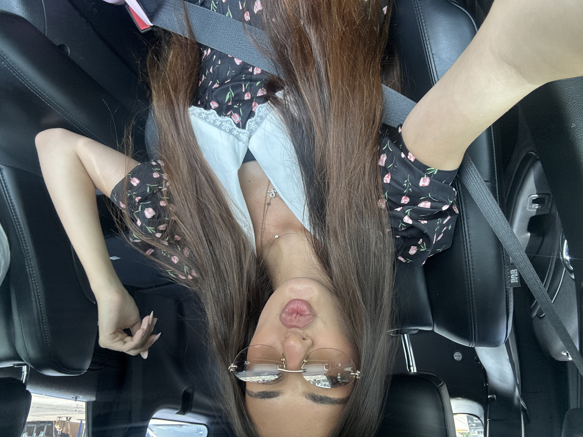A woman with long, brown hair and sunglasses is taking a selfie in the driver's seat of a car. She is wearing a black dress with a floral print and a white collar. She is puckering her lips for the photo. The car's interior is visible, including the black leather seats and a seatbelt. The background shows a blurred outdoor scene, suggesting the car is in a city.