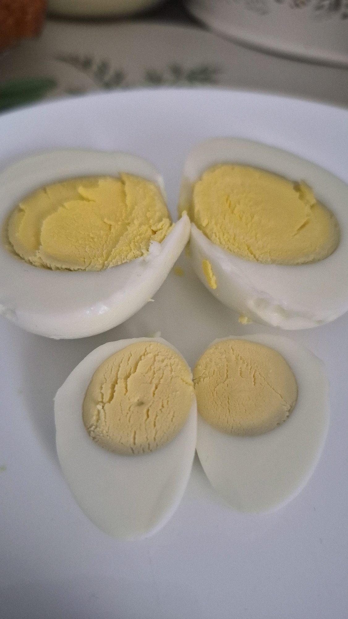 Four halves of hard-boiled eggs are arranged on a white plate. Two larger halves are positioned at the top, with their cut surfaces facing upwards, revealing a yellow yolk. Below these, two smaller halves are similarly arranged. The eggs appear to be cooked to a firm consistency, with the yolks exhibiting a slightly crumbly texture. The lighting is natural, casting soft shadows on the plate. The background is blurred, suggesting an indoor setting, possibly a kitchen or dining area. The image does not contain any elements that would allow for the determination of its location in Krauja, Latvia.