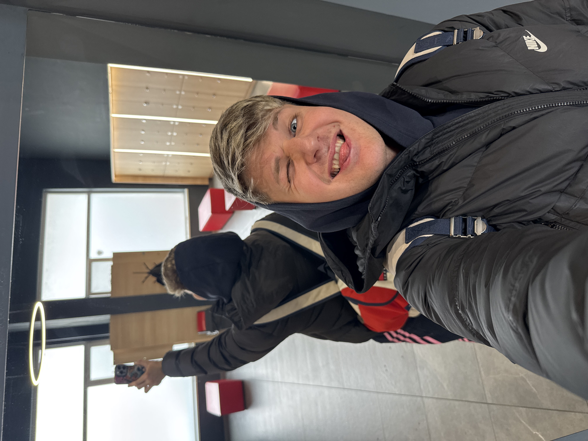 An indoor scene captured as a mirror selfie within a facility in Plovdiv, Bulgaria. In the immediate foreground, a male with light brown hair, wearing a black puffer jacket with a white Nike logo and a dark blue hooded sweatshirt, winks with his right eye, squints his left eye, and protrudes his tongue in a playful expression while holding a smartphone to take the photo. Blue and white backpack straps are visible over his shoulders. In the background, reflected in the same surface, another individual, possibly the same person or a companion, is seen from behind, wearing a dark hooded jacket and a backpack with white straps and a red pouch, holding a smartphone with a colorful case upwards, appearing to photograph or video the surroundings. The environment features dark grey walls, a tiled floor, and large frosted or obscured windows in the background. A luminous circular fixture hangs from the ceiling. To the right, a series of light-colored wooden lockers or cubbies with vertical illumination are visible. Several red, cube-shaped objects are distributed on the floor in the background.