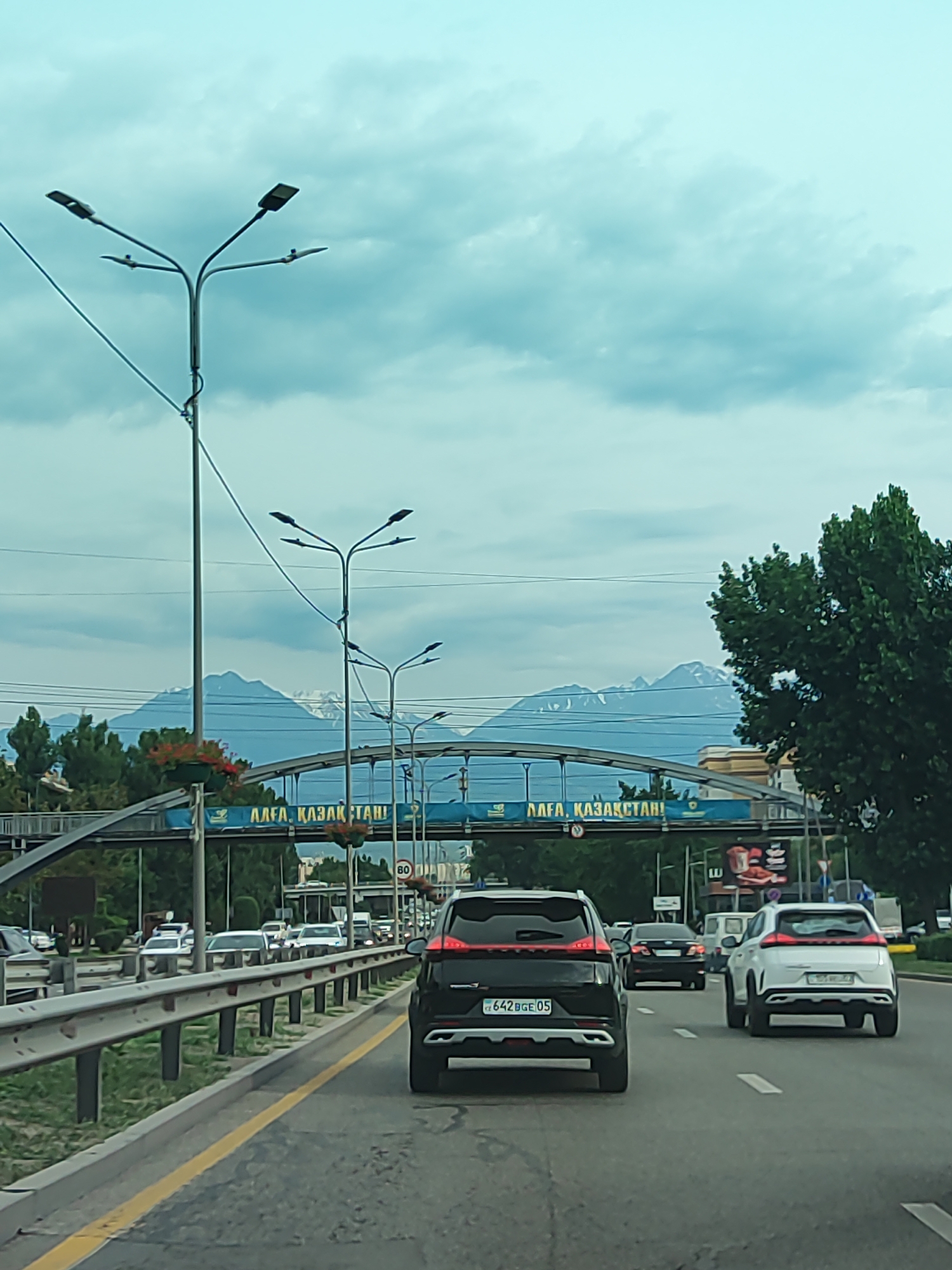 The media file depicts a view from a vehicle driving on a highway, likely within Almaty, Kazakhstan. The scene is set on a multi-lane road with a metal guardrail separating the traffic lanes from the grassy median. In the foreground, several cars are visible, including a black SUV with a license plate ending in "BGE 05" and a white SUV. A pedestrian bridge spans across the highway in the mid-ground, adorned with flowering plants in hanging baskets. Above the bridge, a banner displays text in what appears to be Cyrillic script, reading "АЛҒА, ҚАЗАҚСТАН!" which translates to "Forward, Kazakhstan!". In the background, snow-capped mountains are visible beneath a cloudy sky. The overall atmosphere suggests an overcast day. The presence of streetlights suggests it could be early morning or late afternoon.