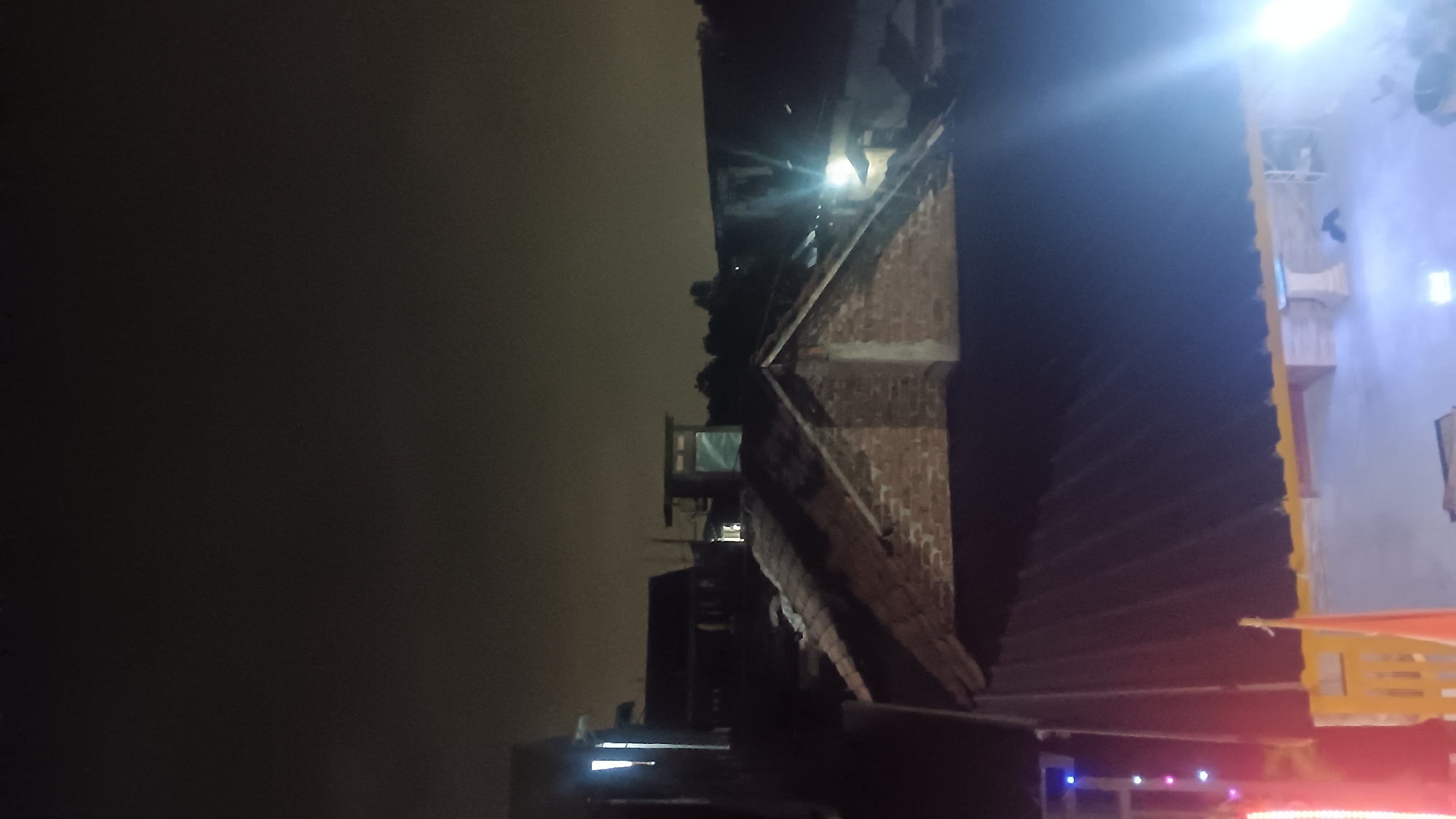 A night-time, slightly overexposed photograph captures a rooftop view in what is identified as the city of Tanjungsari, Indonesia. The scene is dominated by the dark, overcast sky. In the foreground, a dark, corrugated metal roof extends diagonally from the bottom left upwards. Below this roof, a yellow wooden fence and a portion of a concrete structure with some furniture are visible. A flag on a pole, possibly Indonesian given the context, stands on the left side.

Mid-ground features several roofs constructed from terracotta tiles, brickwork, and corrugated metal. Some structures appear to be residential or commercial buildings. Lights are visible in several windows and on some of the structures, casting a glow into the night. One prominent structure in the mid-ground is a small, box-like building with windows, possibly a small office or shelter.

The background consists of a dark tree line and more building silhouettes, suggesting a densely populated area. Bright lights, likely from streetlights or building illumination, are scattered throughout the mid and background, creating lens flares and illuminating parts of the scene. The overall impression is one of a typical urban or semi-urban environment at night. There are no discernible people or animals in the frame.