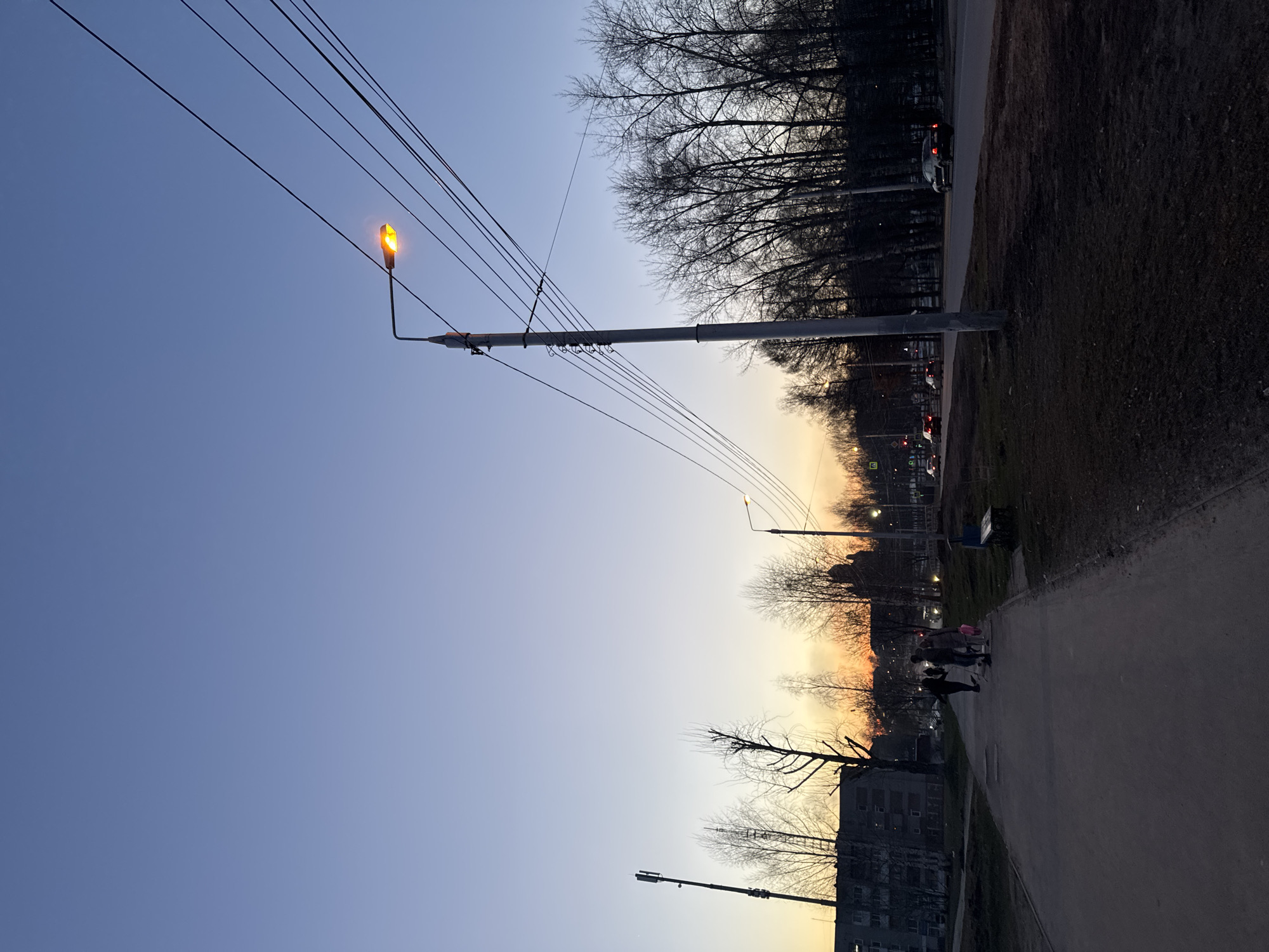 The image depicts a street scene in Berezniki, Russia, at twilight. A paved sidewalk runs along the bottom right, bordering a grassy area. Bare trees line the horizon, silhouetted against a gradient sky transitioning from blue to yellow. Utility poles with multiple electrical wires stretch across the sky, and a lit streetlamp stands prominently. In the distance, figures can be seen walking on the sidewalk. A multi-story building is partially visible on the left, and a car can be seen driving on the road in the background on the right.