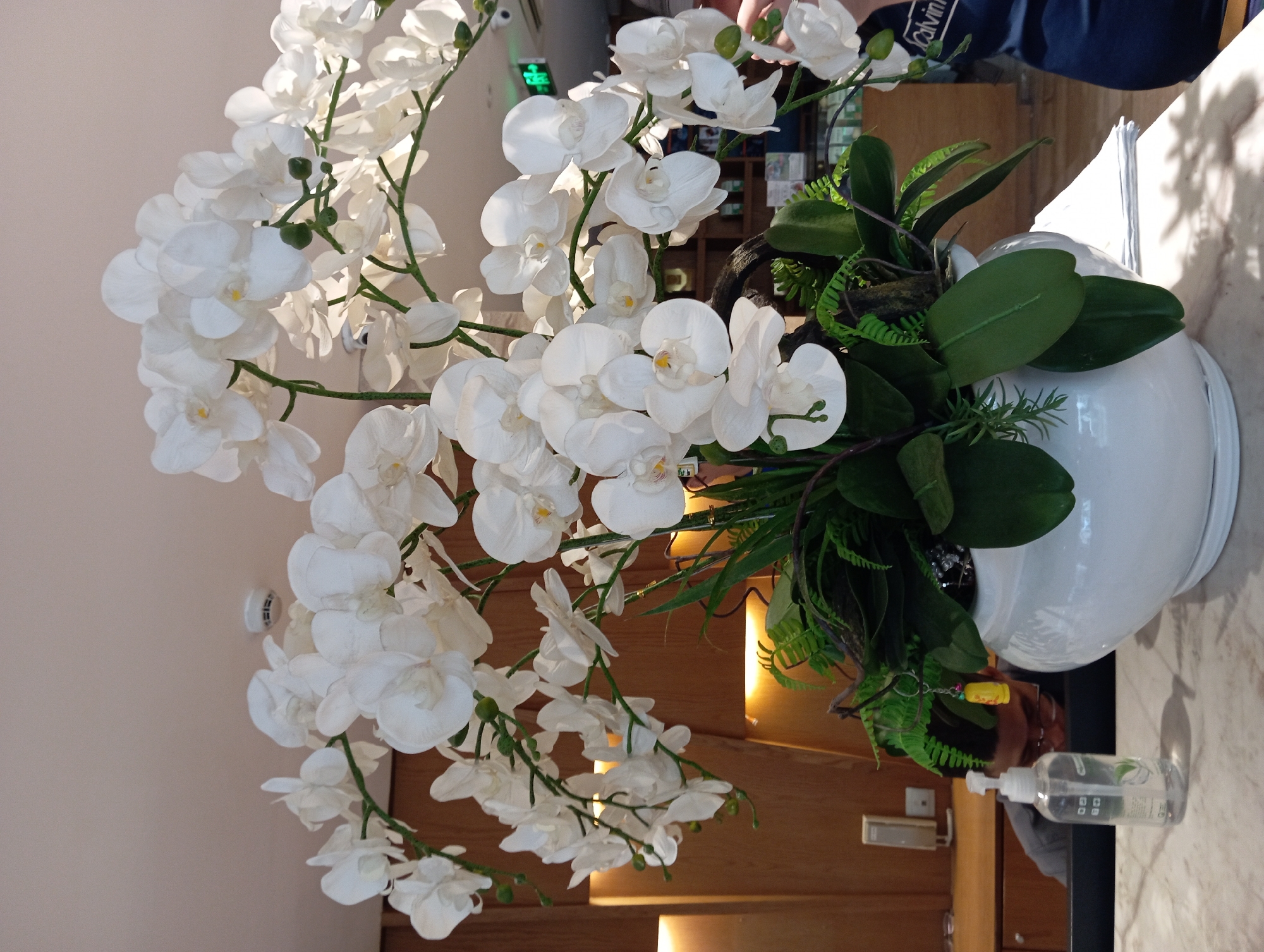 A large, white ceramic pot filled with artificial white orchids and greenery sits on a marble countertop in what appears to be a reception area or lobby. Behind the floral arrangement, a wooden wall with built-in lighting is visible, as is a portion of a counter with a hand sanitizer dispenser. In the background, a person wearing glasses is partially visible, appearing to be working at a desk. A security camera is mounted on the ceiling, and a green exit sign is visible on the wall. The overall impression is of a well-maintained indoor space, possibly a hotel or office. The image does not contain enough information to ascertain the specific location within Nha Trang, Vietnam.