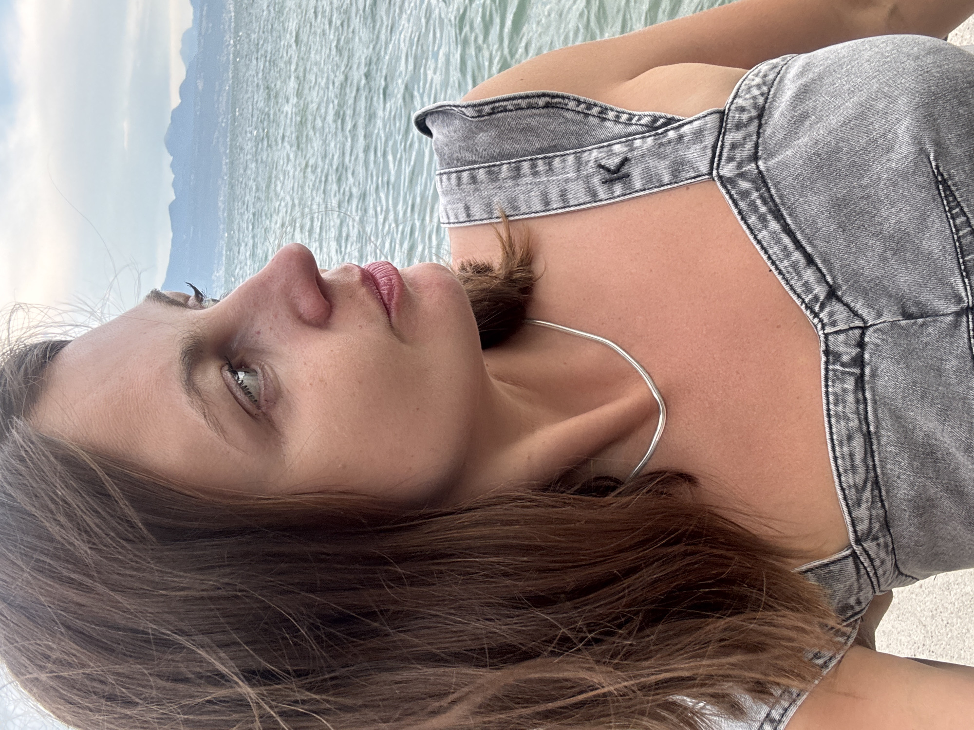 The image is a medium shot of a woman from the chest up, taken from a side profile. She has brown hair, fair skin, and is looking to her right, out towards a body of water. She is wearing a gray denim top with thin straps. Around her neck, she has a thin silver necklace. In the background, there is a large body of water, likely Lake Geneva, and a hazy mountain range in the distance, which is characteristic of the landscape near Lausanne, Switzerland. The sky is overcast with clouds. The woman appears to be outdoors, possibly on a pier or a waterfront promenade.