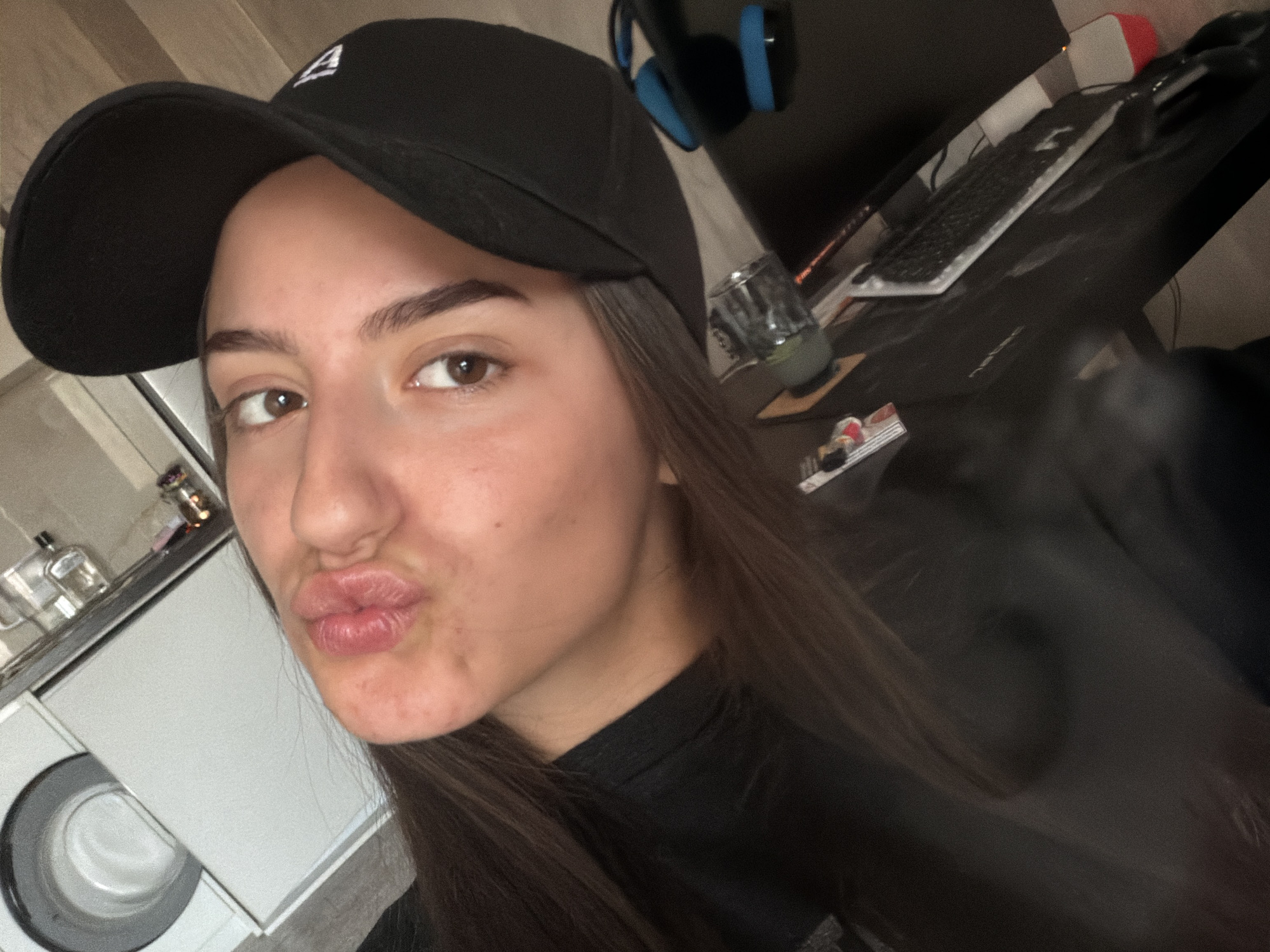A young woman with brown hair, wearing a black t-shirt and a black baseball cap, is taking a selfie. She is puckering her lips and looking at the camera. She appears to be indoors, in what looks like a domestic setting. In the background, there is a washing machine and a countertop with various items on it. To her right, a desk with a computer keyboard and headphones is visible. The lighting is dim and appears to be artificial. There are no visible indicators of the location being in the city of Burgas, Bulgaria.