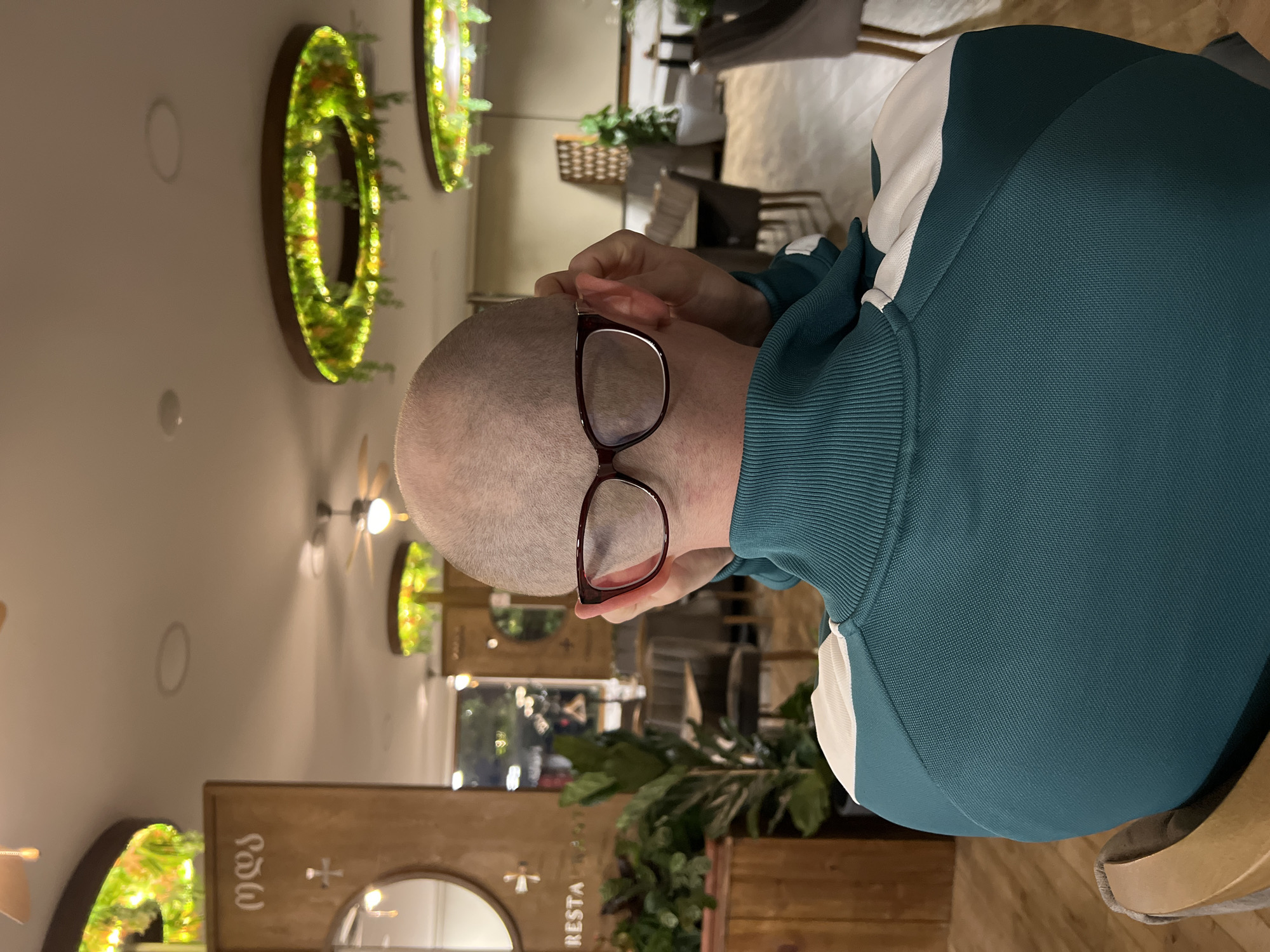 The image shows a person with a shaved head, wearing glasses and a teal-colored sweatshirt with a white stripe. The person is adjusting their glasses with their hand. They appear to be sitting in a restaurant or cafe. The background includes circular decorations with greenery on the wall, as well as other tables and chairs. There are plants in wooden boxes and a wooden sign that reads "RESTA". The location is identified as Boreti, Montenegro.