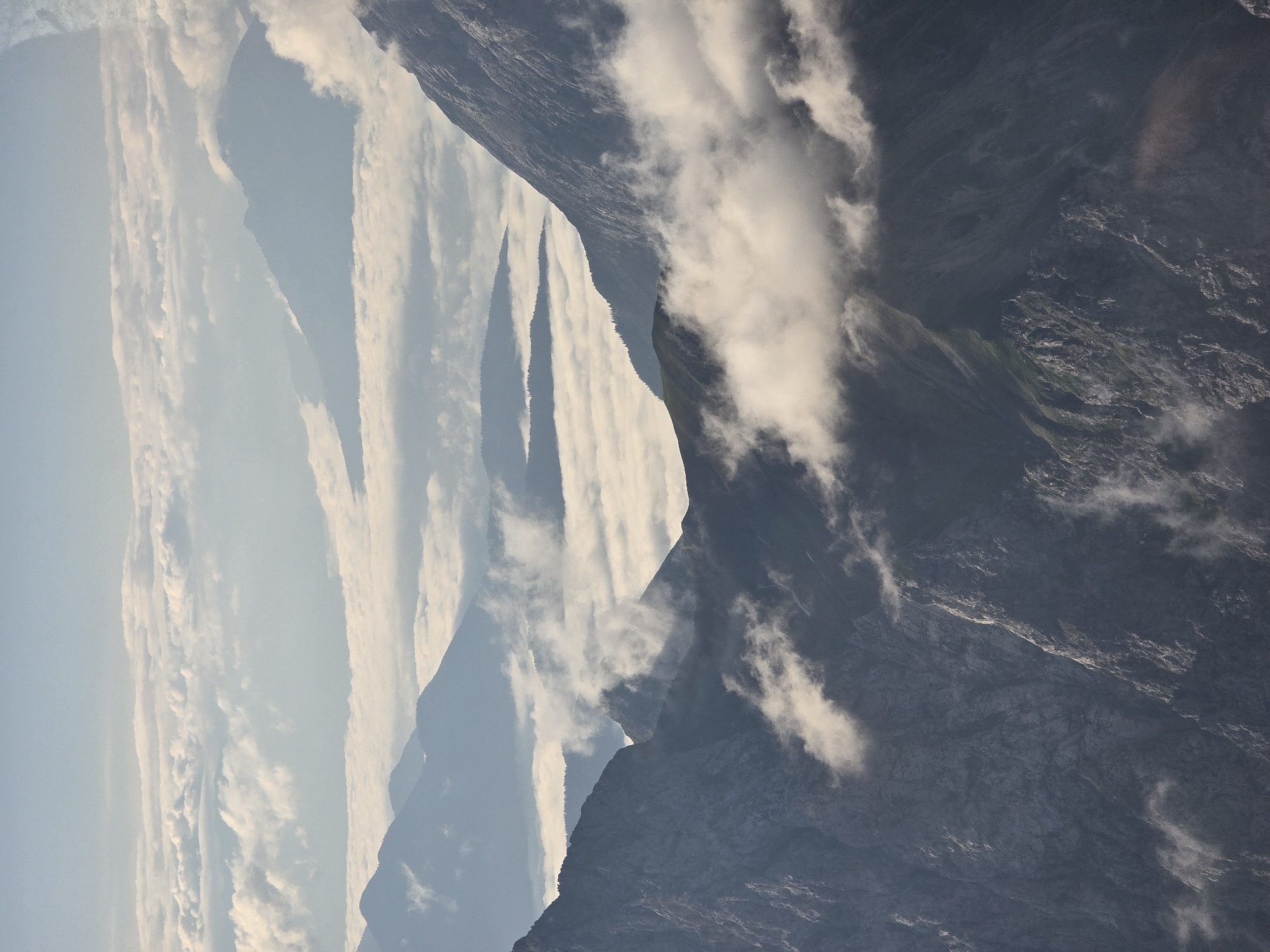 This aerial image depicts a mountainous landscape covered in a sea of clouds, as seen from a high vantage point, likely above the tree line. The foreground is dominated by dark, rugged mountain peaks with visible rocky textures. Wisps and patches of white clouds are scattered amongst these peaks, obscuring some of the lower slopes and adding depth to the scene.
Beyond the immediate foreground, layers of mountains recede into the distance, their silhouettes softened by atmospheric haze and the pervasive cloud cover. These further mountain ranges appear as muted blue and gray shapes, some with visible peaks emerging from the cloud layer. The clouds create a distinct division between the upper and lower portions of the landscape, giving the impression of an ocean of mist.
The sky above is a pale blue, transitioning to a lighter hue towards the horizon, with subtle hints of clouds. The lighting suggests it is either early morning or late afternoon, with soft, diffused sunlight illuminating the scene.
The image captures the natural grandeur of the Bavarian Alps, a region that includes Garmisch-Partenkirchen. While no specific structures or human presence are visible, the scene evokes the remote and majestic character of the alpine environment.
