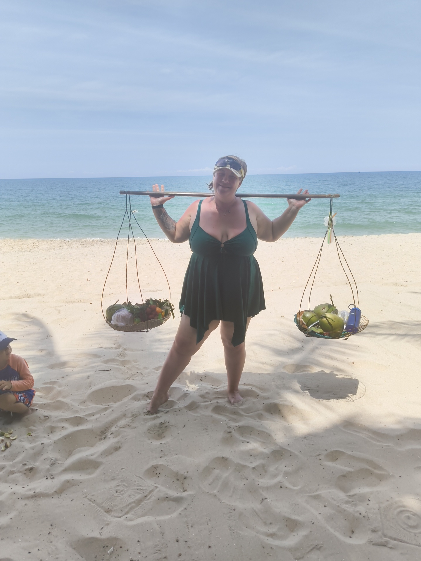 A full-body, eye-level, outdoor shot captures a woman standing on a sandy beach. She is wearing a dark green swimsuit with a v-neck and a draped, asymmetrical skirt. She has a bandana on her head and sunglasses on her forehead. The woman is carrying a wooden yoke across her shoulders, with two baskets of produce hanging from it. The left basket contains an assortment of fruits, including red and yellow items, and a white wrapped object. The right basket holds several green coconuts and a blue thermos. Her arms are outstretched, supporting the yoke. To the left of the woman, a child wearing an orange and purple rashguard and patterned shorts is seated on the sand, looking down. The background consists of a tranquil blue ocean meeting a pale blue sky with scattered clouds. The sand is a light beige color, with visible footprints and shadows cast from unseen objects. The scene is set in Phu Thuy Ward, Vietnam.