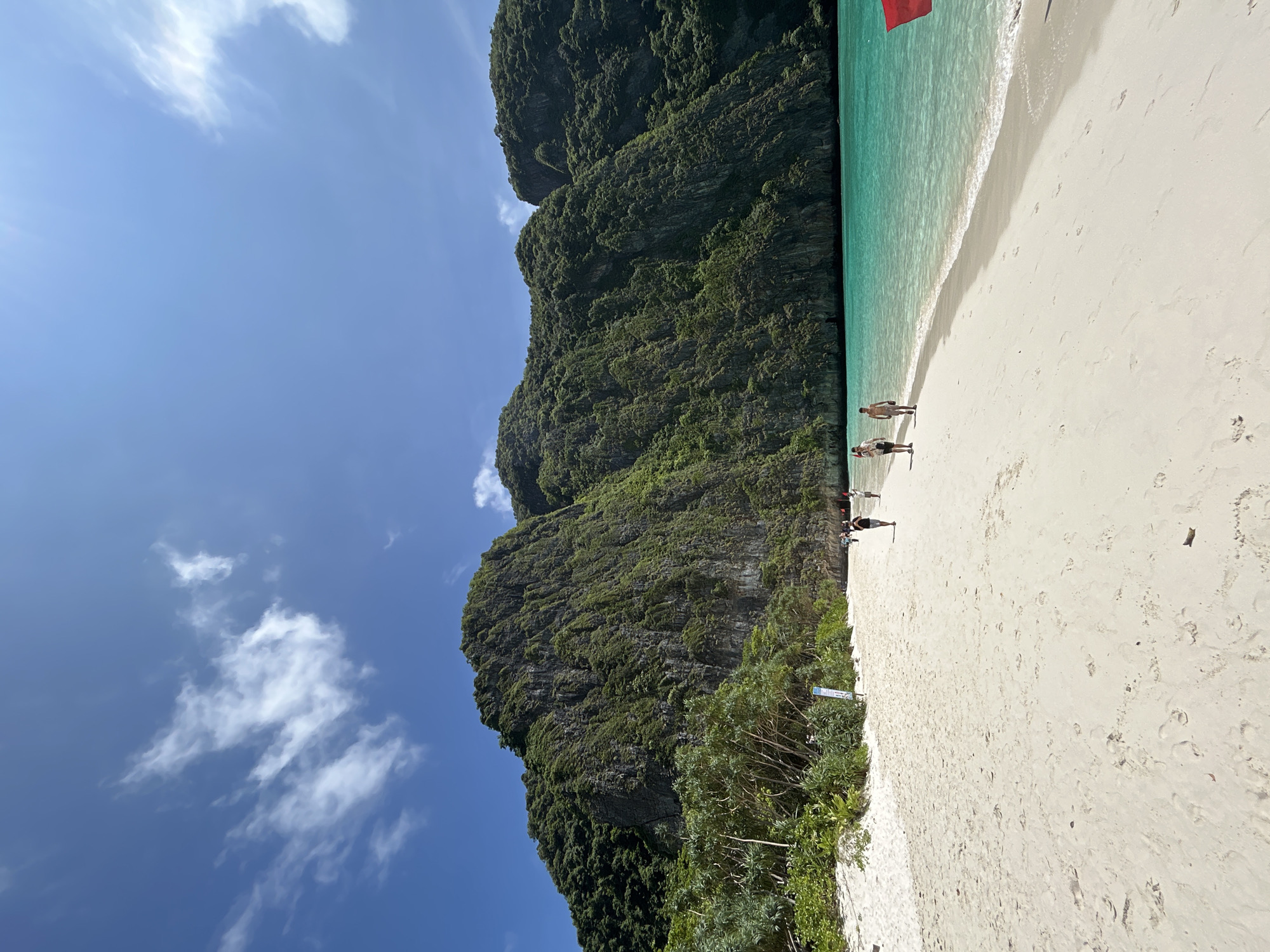 The image depicts a tropical beach scene with clear turquoise waters, a white sandy shore, and lush green cliffs in the background. The sky is blue with scattered white clouds. Several people are walking along the beach, some wading in the shallow water. The location is likely Maya Bay, Krabi, Thailand, which is a famous tourist destination known for its stunning natural beauty.