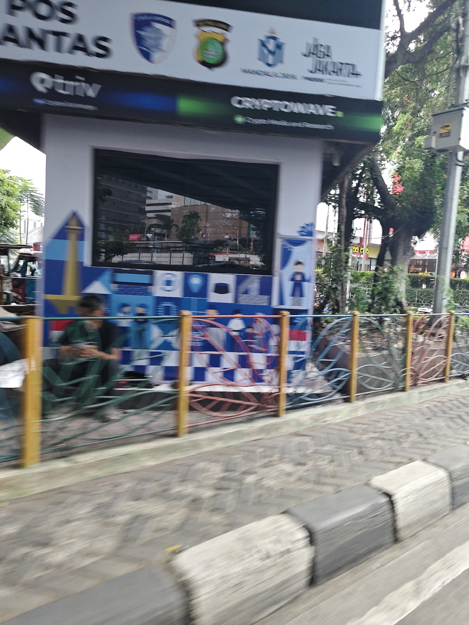 The image shows a blurry view of a street scene in West Jakarta, Indonesia. In the foreground, there is a curb with alternating black and white sections. Beyond the curb, there is a road surface that is also out of focus. In the background, a white structure with a large window is visible. The structure appears to be a kiosk or small building, possibly a public service point, as suggested by the text "POS ANTAS" which likely refers to a police station or public service center. The window reflects buildings and trees. The structure is decorated with colorful geometric patterns, including blue and white squares and a depiction of the Monas tower. There are also logos and text on the structure, including "Qtr iv" and "CRYPTOWAVE". Several people are visible near the structure, some sitting on the ground. The overall scene is blurry due to motion, suggesting the photo was taken from a moving vehicle. Trees and foliage are visible in the background, indicating an urban environment with greenery.