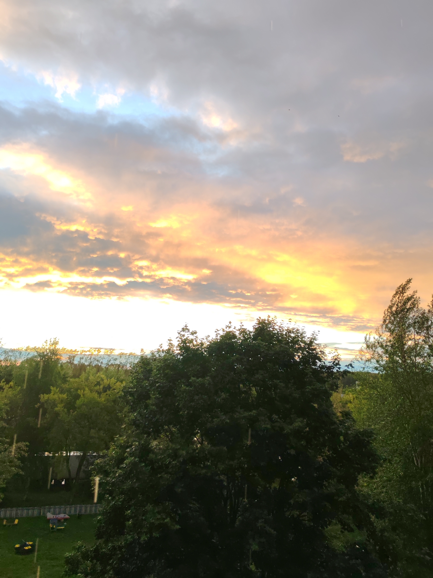 This is a medium shot, eye-level photograph of a park-like setting with a dramatic sky. The main subjects are the lush green trees in the foreground and midground, with a prominent, large, dark green tree dominating the center and right of the frame. Behind the trees, the sky is filled with colorful clouds illuminated by the setting sun, casting a warm, golden-orange glow across the scene. The sky transitions from these warm hues to cooler blues and grays at the top. Streaks of rain are visible, indicating a light shower.
In the background, a fence surrounds a grassy area with some playground equipment, including what appears to be a small slide or play structure with red and gray components, and yellow benches or seats. A yellow lawnmower or similar equipment is also visible on the grass. The presence of a fenced-in play area and well-maintained trees suggests a residential park or backyard in a suburban or urban environment.
The time of day is clearly sunset, given the vibrant colors in the sky. The weather is a mix of rain and sunshine, creating a potentially moody or picturesque atmosphere. The overall emotion conveyed is one of natural beauty and perhaps tranquility, despite the presence of rain. There are no visible text elements in the image. The location is identified as Сокол, Russia.