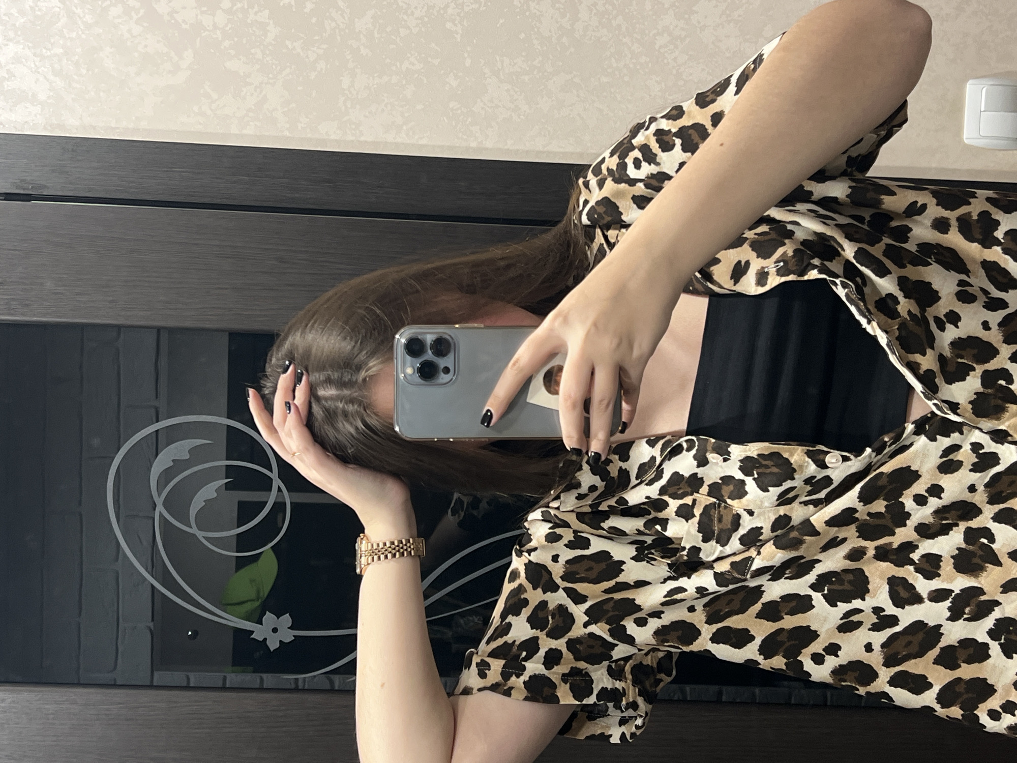 A person takes a selfie indoors, holding a gray smartphone. The person is wearing a black tank top and a leopard-print shirt. The person has a gold watch on their wrist. Behind the person is a dark gray door or cabinet with a decorative swirl design on it. The background is a beige wall. The photo may have been taken in Chaikovsky, Russia.