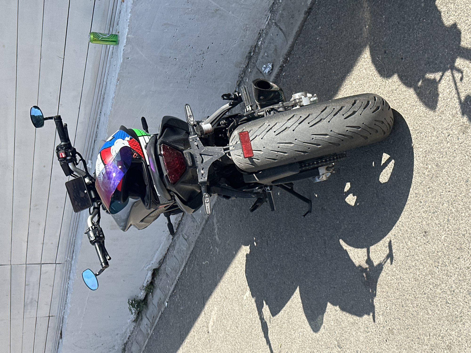 A parked motorcycle is captured from a rear-facing perspective, angled slightly to the left. The motorcycle is dark in color and has a custom helmet resting on its seat, featuring a vibrant comic-book-style graphic. In the background, a white concrete wall with horizontal indentations runs along the scene. To the right, on top of the wall, sits a green soda can. The motorcycle's license plate is visible, and its rear tire is prominent. A shadow of the motorcycle stretches across the asphalt ground, indicating a sunny day. The setting appears to be a street in Budva, Montenegro, suggested by the architecture and general environment.