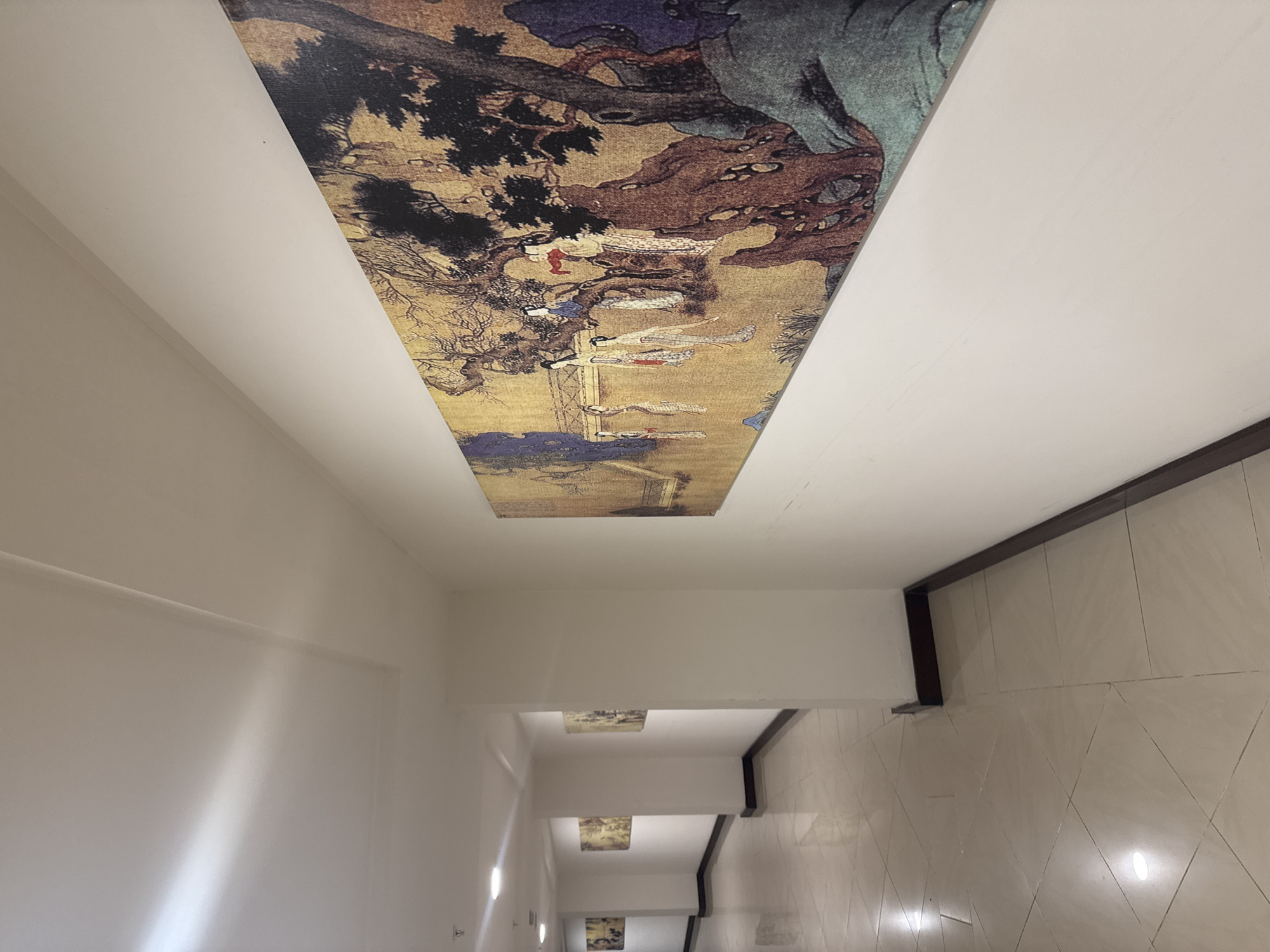The image depicts a modern, well-lit interior hallway with a polished tiled floor. The hallway features a series of framed artworks displayed on the walls. The artwork prominently featured in the foreground is a long, horizontal painting with a golden background, depicting traditional East Asian figures in a naturalistic setting with trees and rocks. Several similar artworks are visible further down the hallway on both sides, suggesting an art gallery or exhibition space. The architecture of the hallway includes white walls, a drop ceiling with recessed lighting, and dark trim along the baseboards. The overall impression is one of an organized and aesthetically curated indoor environment. The specific location in Aksu, Turkey, cannot be determined from the visual information provided in the image.
