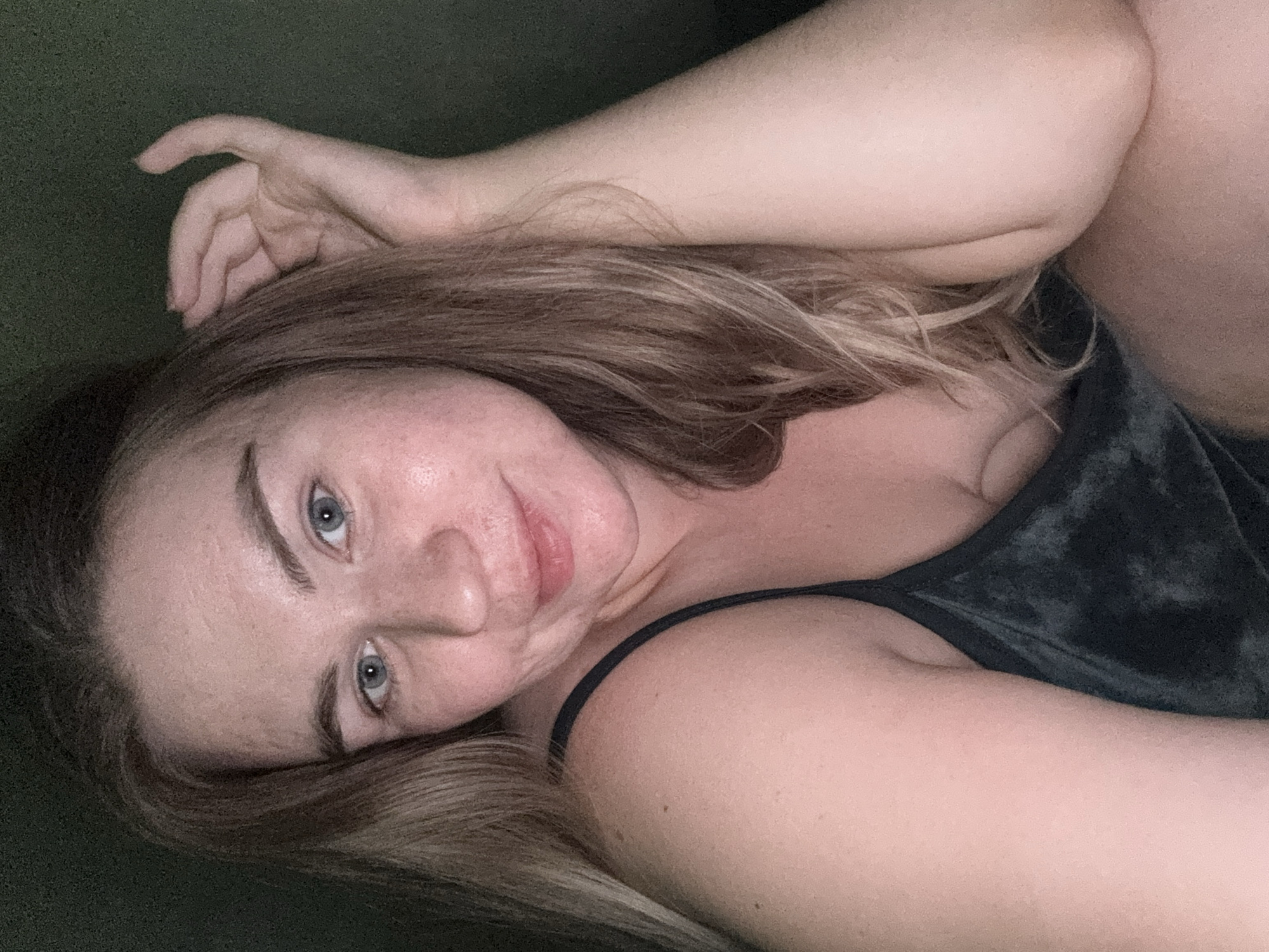 A woman with light blonde hair and blue eyes is lying down, possibly on a dark green surface like a couch. She is wearing a dark-colored tank top. Her left arm is raised with her hand near her head. The image is cropped and does not provide information about location in Chaikovsky, Russia or any specifics of the environment.
