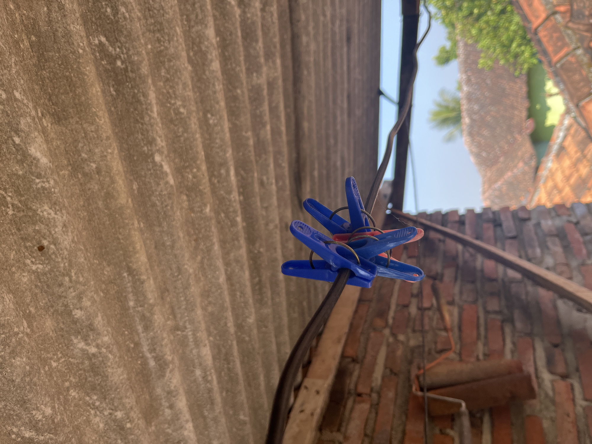 A close-up, low-angle outdoor shot captures a group of blue clothespins clamped onto a black wire that stretches horizontally across the frame. The clothespins are clustered together and appear to be holding something, though the object is not visible. Behind the clothespins and wire, a brick wall forms a textured backdrop, with some wooden beams visible. Above this, a corrugated, weathered roof runs diagonally from the top left to the bottom right. In the background, out-of-focus houses with tiled roofs and lush green trees suggest a residential area in Glagah Kulon, Indonesia, under a bright, clear sky. The lighting indicates it is daytime.