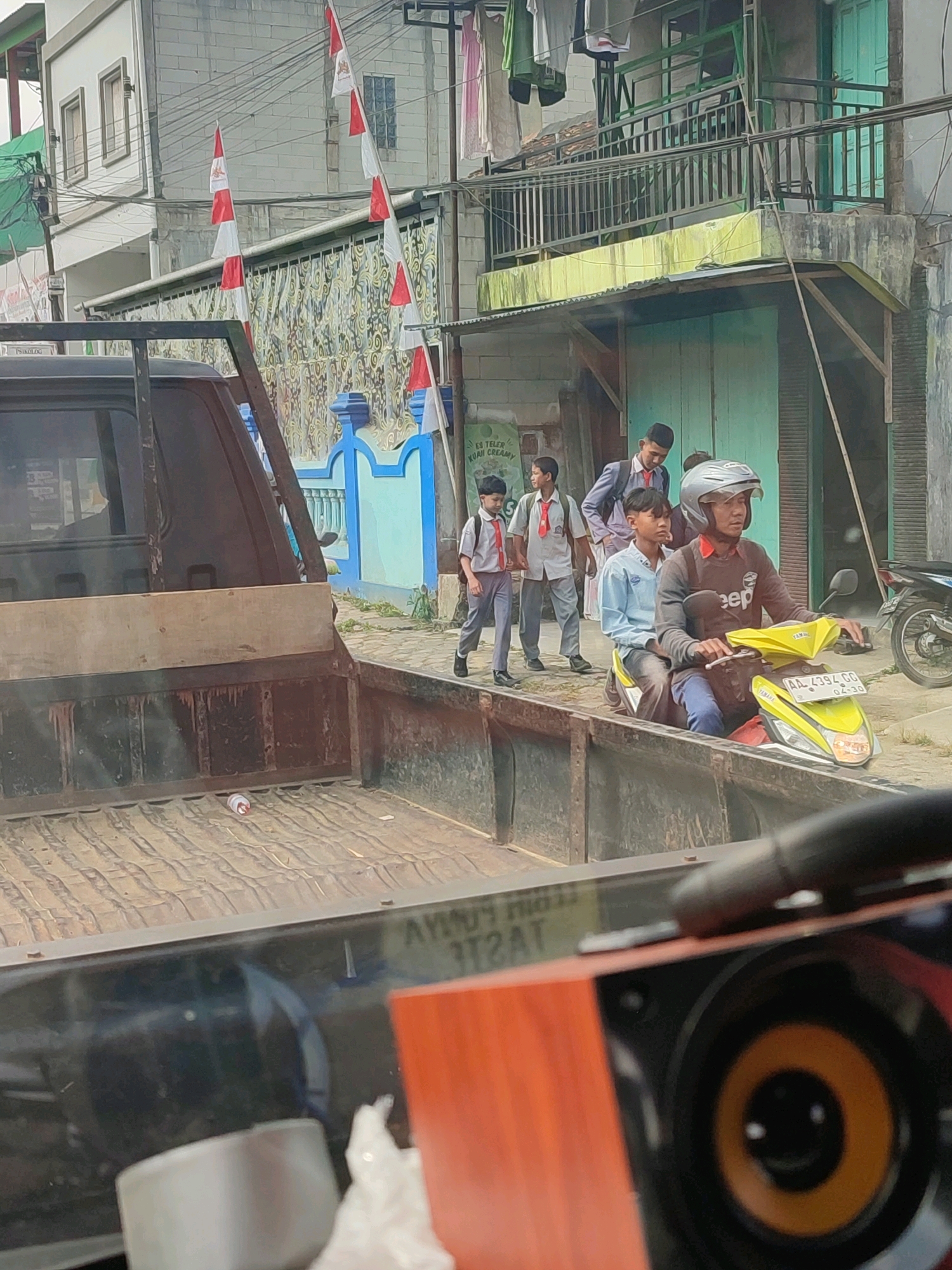 In Kalikajar, Indonesia, a scene unfolds on a street lined with buildings and flags. In the foreground, the bed of a pickup truck is visible, with a white and red bottle lying on its floor. A group of four young males, seemingly students wearing school uniforms and red ties, are walking on the sidewalk. One of them is being interviewed. Behind them, two adult males are riding a yellow scooter. The scooter rider is wearing a helmet and a dark t-shirt with the word "Jeep" on it. The scooter has a license plate that reads "AA 4394 GC". The background shows a building with balconies and laundry hanging out to dry, as well as more flags and a decorative wall. The overall impression is a snapshot of everyday life in a residential area, possibly during a national holiday given the prominent display of flags.