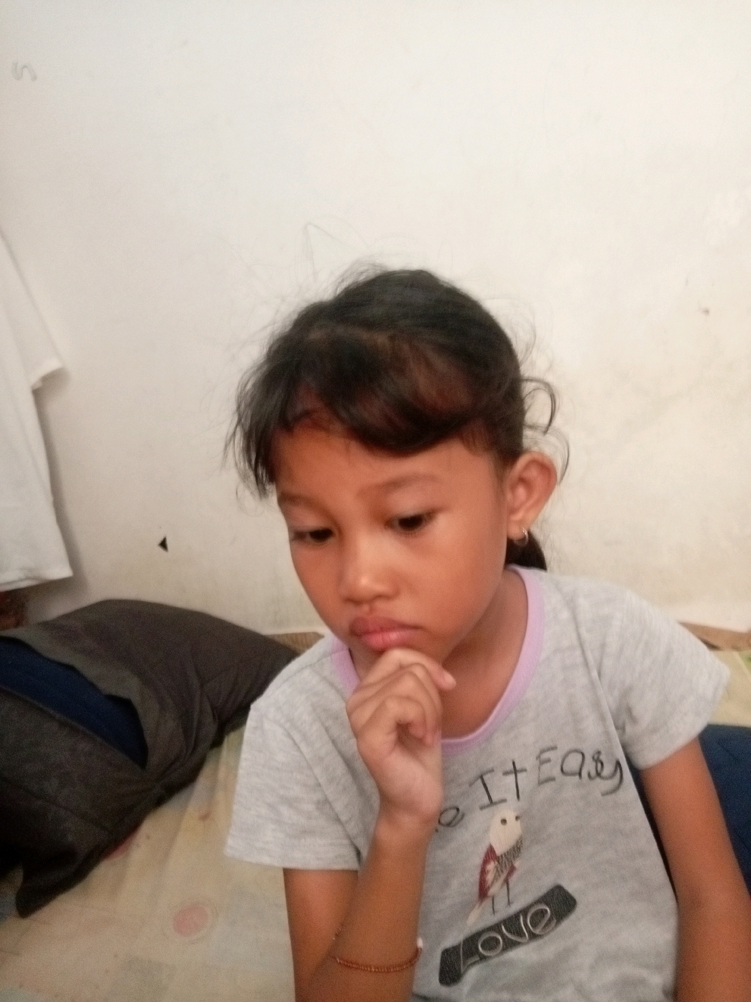 A young Indonesian girl with dark hair and brown eyes is shown in a close-up shot, looking down and resting her chin on her hand. She is wearing a light gray t-shirt with a graphic design featuring a bird and the words "Do It Easy" and "Love" written on it. The t-shirt has a light purple trim around the neckline. The girl has a small hoop earring in her left ear. She is positioned in the foreground, with a blurred background that includes a wall and what appears to be a dark gray pillow or cushion. The lighting suggests an indoor setting, possibly a home. The overall tone of the image is candid and thoughtful. The location in Batam, Indonesia, is not explicitly evident from the visual cues within the image itself.