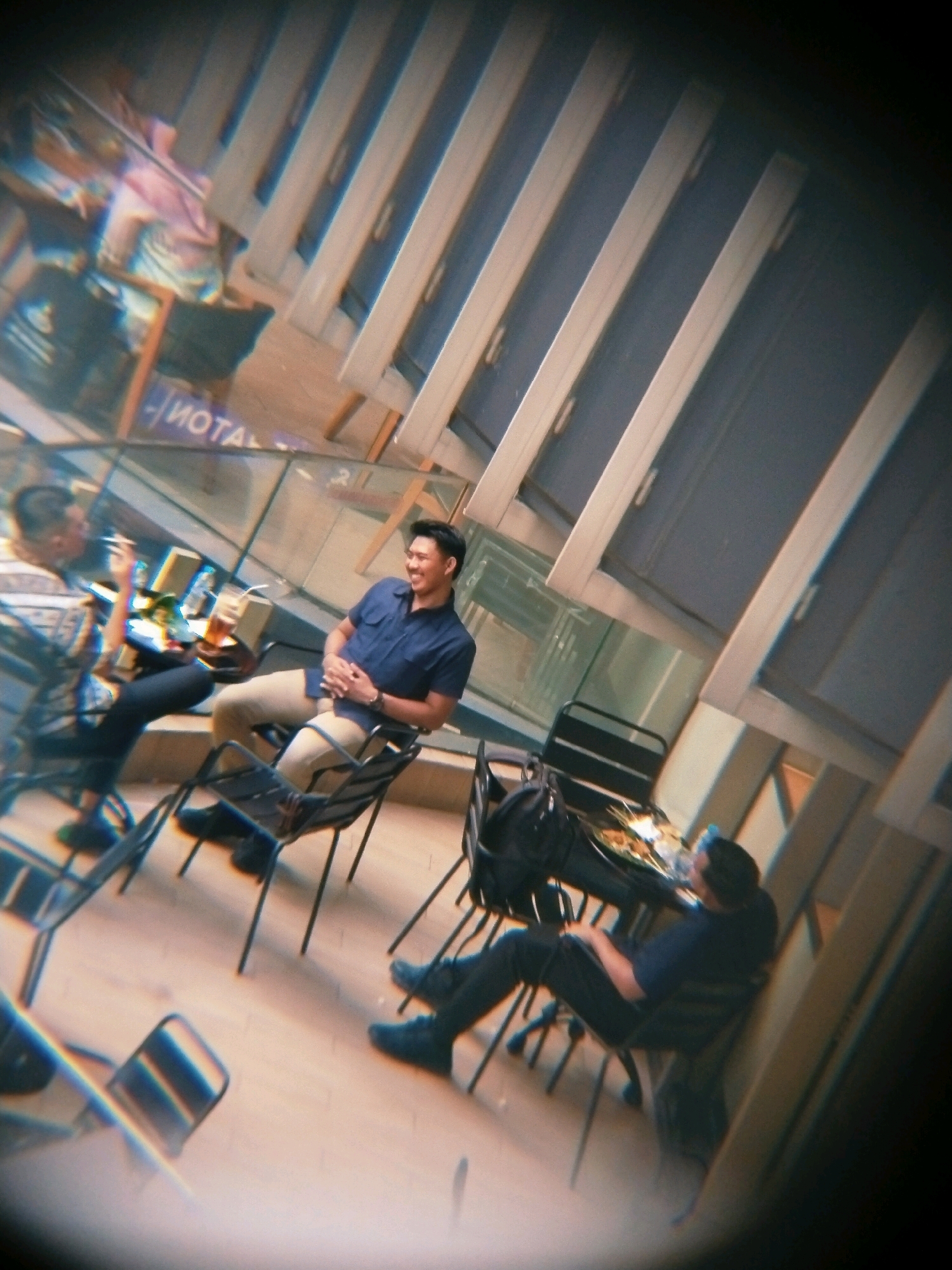 The image depicts an indoor scene, likely a cafe or restaurant, with a distorted, vignetted effect. In the foreground, a man in a blue collared shirt and tan pants is seated at a black metal chair, facing right and smiling. His hands are clasped in his lap. To his left, another man in a patterned shirt and dark pants is seated, holding a cigarette in his right hand, and appears to be looking down. A table with drinks and food is between them. Further in the background, a third person is seated, with their legs crossed and wearing dark pants and shoes. The environment features wooden flooring, and the background has a series of parallel white beams and dark panels, possibly windows or architectural elements. A sign with the text "MOTA" is partially visible on a glass partition. The overall lighting is dim. The presence of the "MOTA" sign suggests a possible connection to the city of Surabaya, Indonesia, as it is a known establishment there.
