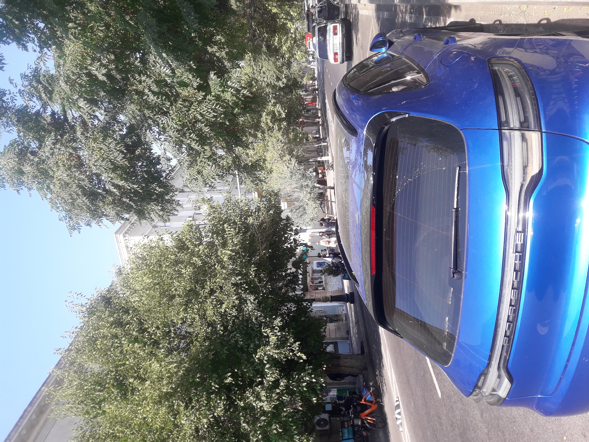A blue Porsche SUV is parked on the side of a street in an urban environment, with its rear facing the camera. Trees with green foliage line the street, casting shadows. Buildings are visible in the background. There are other cars and a bicycle on the street. People are walking on the sidewalk. The sky is clear and blue.