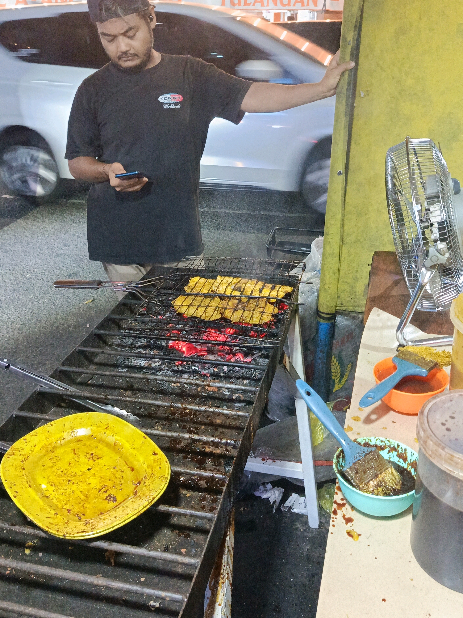 A street food vendor stands by a charcoal grill, wearing a black t-shirt and a cap, while looking at his phone. The grill is laden with pieces of yellow-marinated food cooking over glowing red coals. To the left, a yellow plate sits on the grill. To the right, a metal fan is positioned near a table with a bowl of sauce and a brush, along with a plastic container. The scene appears to be a roadside stall in Tangerang, Indonesia, indicated by the text "TULANGAN" visible on a sign in the background, which is a common term for bone-in meat dishes in Indonesian cuisine, and a glimpse of a white vehicle, possibly a car, passing by. The overall impression is of a bustling evening street food operation.