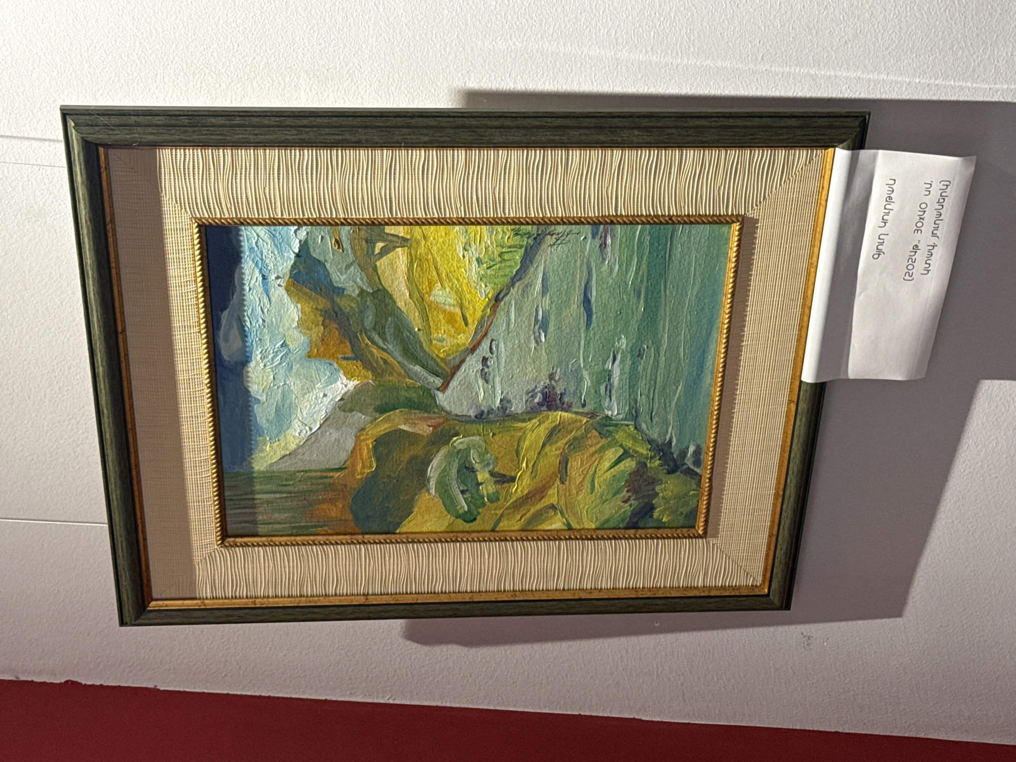 A framed impressionist painting hangs on a white wall, with a red surface visible in the lower left. The painting features bold brushstrokes in shades of blue, green, and yellow. A beige textured mat surrounds the painting, and a dark green frame encases the mat. A small white card is affixed to the frame's right side. The location is the city of Yerevan, Armenia, inferred from the language on the card.