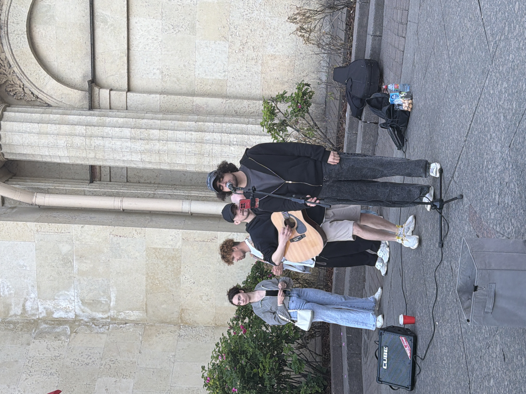 The image depicts a group of street performers in Saint Petersburg, Russia, positioned in front of a building with classical architecture featuring large columns and a stone facade. A young man wearing a black hoodie and a cap is the primary vocalist, singing into a microphone. To his left, another young man is playing an acoustic guitar. A woman in a grey jacket and light-colored jeans stands to the far left, observing the performance. Another person, partially visible, is positioned behind the guitarist.
On the ground, various objects are scattered, indicating the performers' setup: a black amplifier labeled "CUBE", a red paper cup, various bags and cases including a guitar case, and a grey storage bin. The ground is paved with cobblestones, and a low stone wall with steps is visible in the background. The scene appears to be set during daytime.