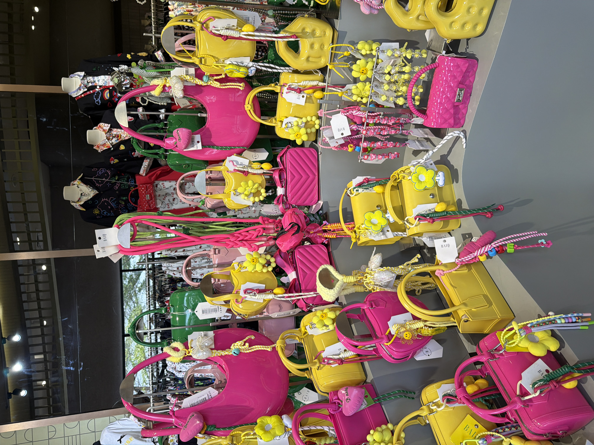 The image displays a vibrant assortment of handbags and accessories displayed on shelves in a retail setting. The items are predominantly in bright, saturated colors, including hot pink, yellow, and green. Many of the handbags are small to medium-sized and feature glossy finishes. Several are embellished with charms, such as flowers, smiley faces, and pom-poms, along with beaded keychains. The background suggests a store interior with clothing racks and mannequins visible, some wearing black tops adorned with playful graphics. The overall aesthetic is cheerful and youthful. The location is specified as a store in East Jakarta, Indonesia.