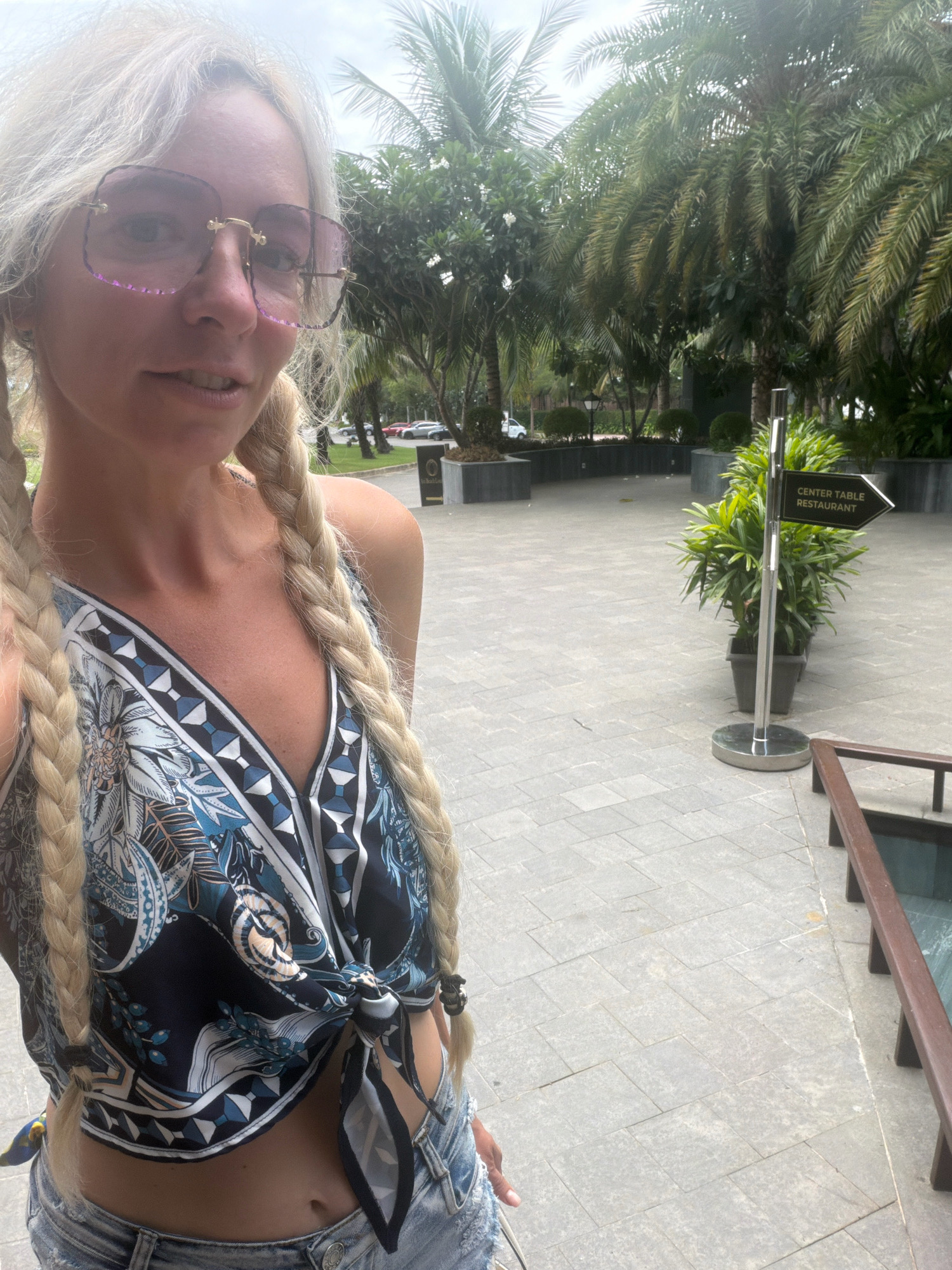 A blonde woman with pigtails and pink-tinted square glasses is taking a selfie outdoors in Cam Lâm, Vietnam. She is wearing a patterned cropped top tied at the waist and denim shorts. Behind her, palm trees and lush greenery are visible, as well as a paved walkway and planters. A directional sign pointing to "CENTER TABLE RESTAURANT" is located to her right. The sky appears overcast.